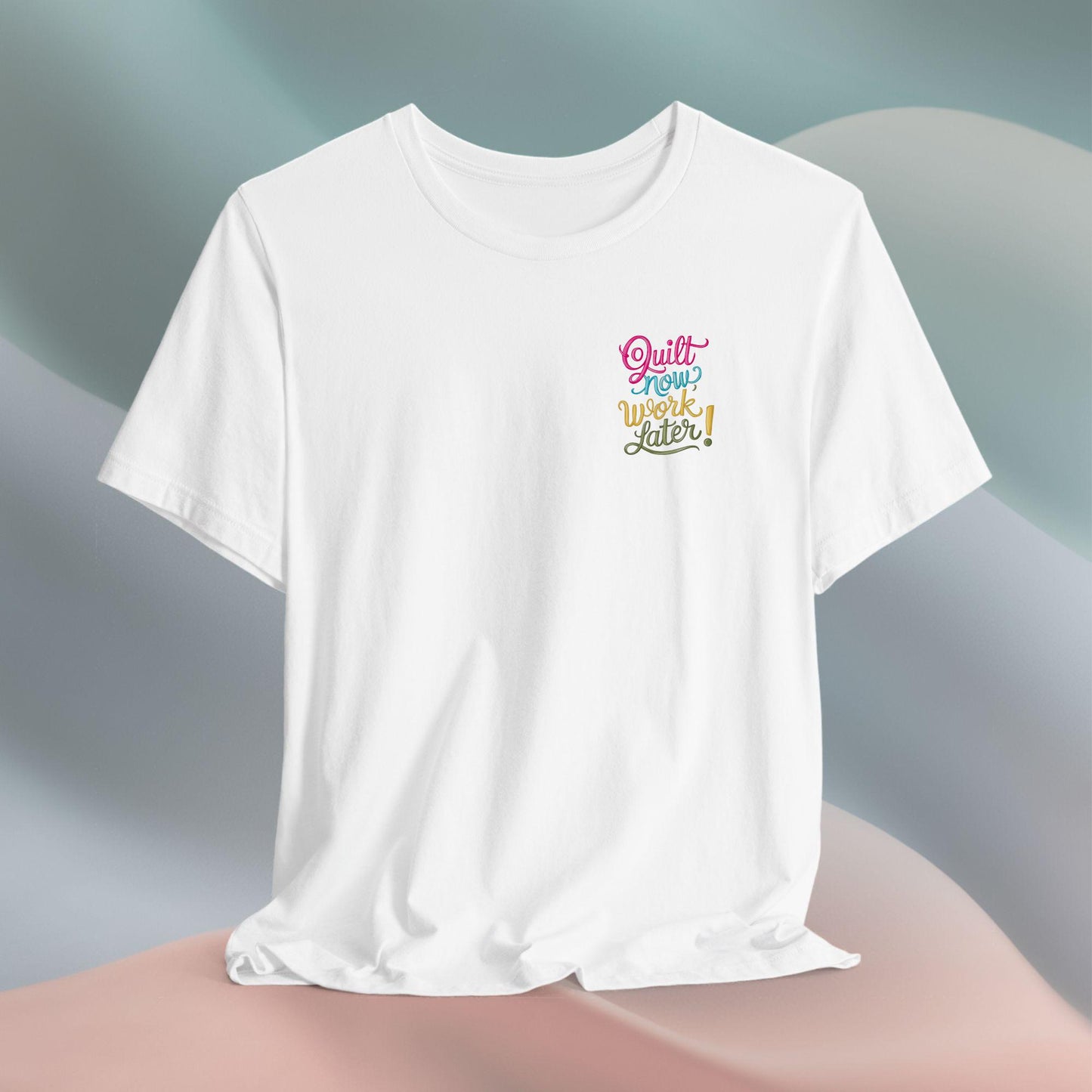 A funny quilting T-shirt featuring bold, stylized text that reads "Quilt Now, Work Later" in various vibrant colors. The playful design captures the spirit of passionate quilters who prioritize their craft over other responsibilities.