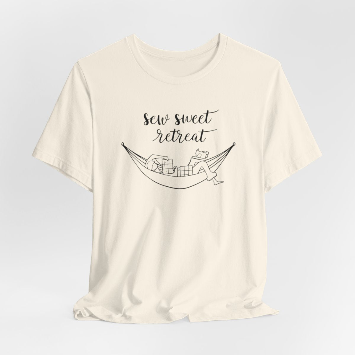 A Natural T-Shirt with a black and white illustration of a hammock filled with quilting supplies like fabric squares and a sewing machine. Text above reads "Sew Sweet Retreat" in a flowing script font.