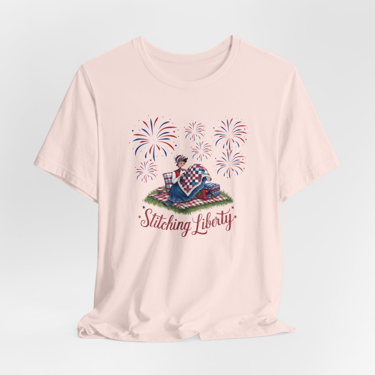 A Soft Pink T-shirt with illustrated scene of a colonial woman quilting a patriotic blanket, surrounded by fireworks. Text reads "Stitching Liberty" in a decorative font.