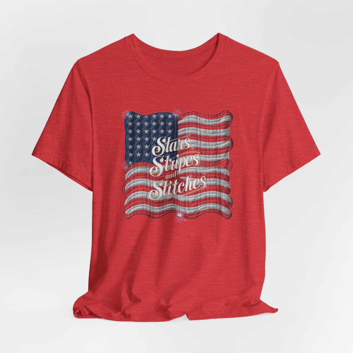 A Heather Red T-shirt featuring quilted American flag design with text "Stars Stripes and Stitches" overlaid in stylized, patriotic font. Fireworks accent the corners.