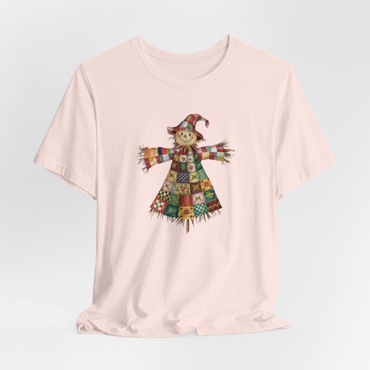 A Soft Pink thanksgiving  quilting t-shirt featuring a smiling scarecrow made entirely of colorful quilt patterns. Wearing a patchwork hat and clothes with various quilt designs. Straw details on hands and neck. Cheerful autumn-themed illustration celebrating quilting craft.