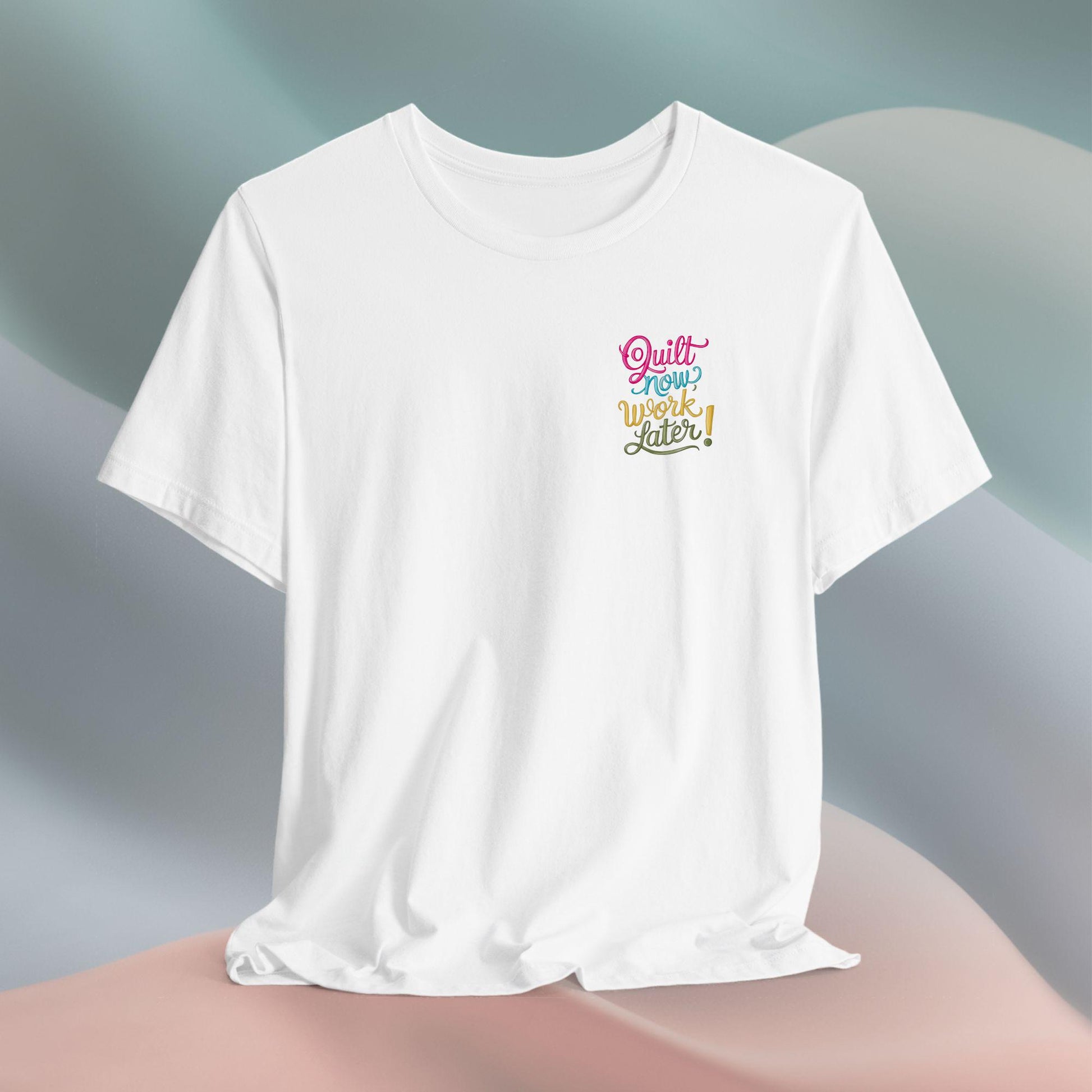 A funny quilting T-shirt featuring bold, stylized text that reads "Quilt Now, Work Later" in various vibrant colors. The playful design captures the spirit of passionate quilters who prioritize their craft over other responsibilities.