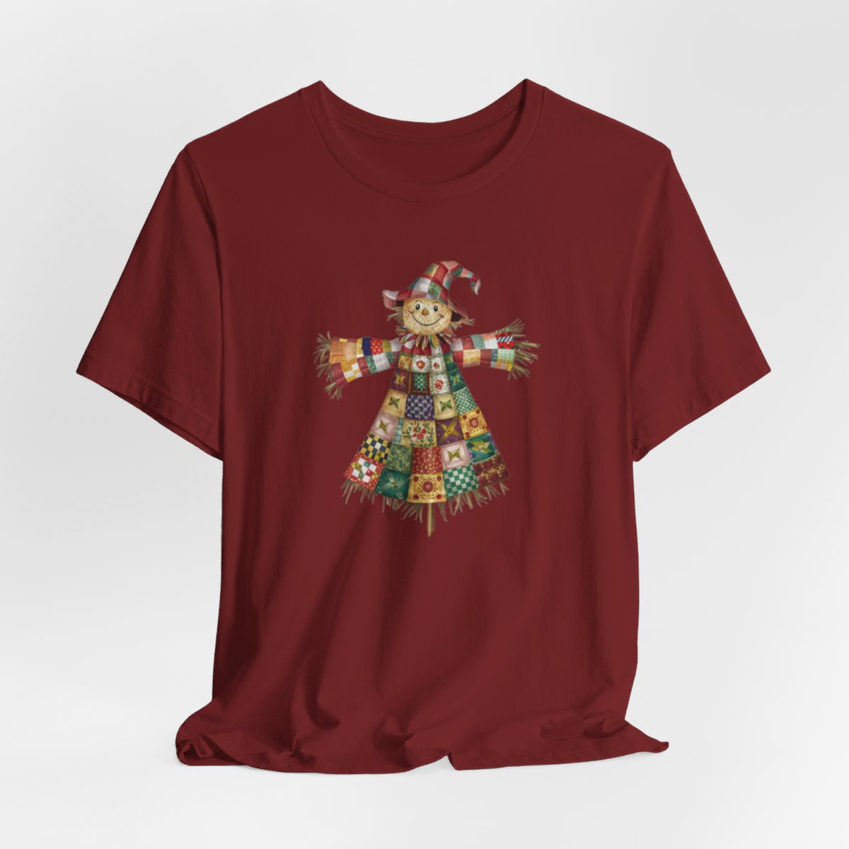 A Cardinal thanksgiving  quilting t-shirt featuring a smiling scarecrow made entirely of colorful quilt patterns. Wearing a patchwork hat and clothes with various quilt designs. Straw details on hands and neck. Cheerful autumn-themed illustration celebrating quilting craft.