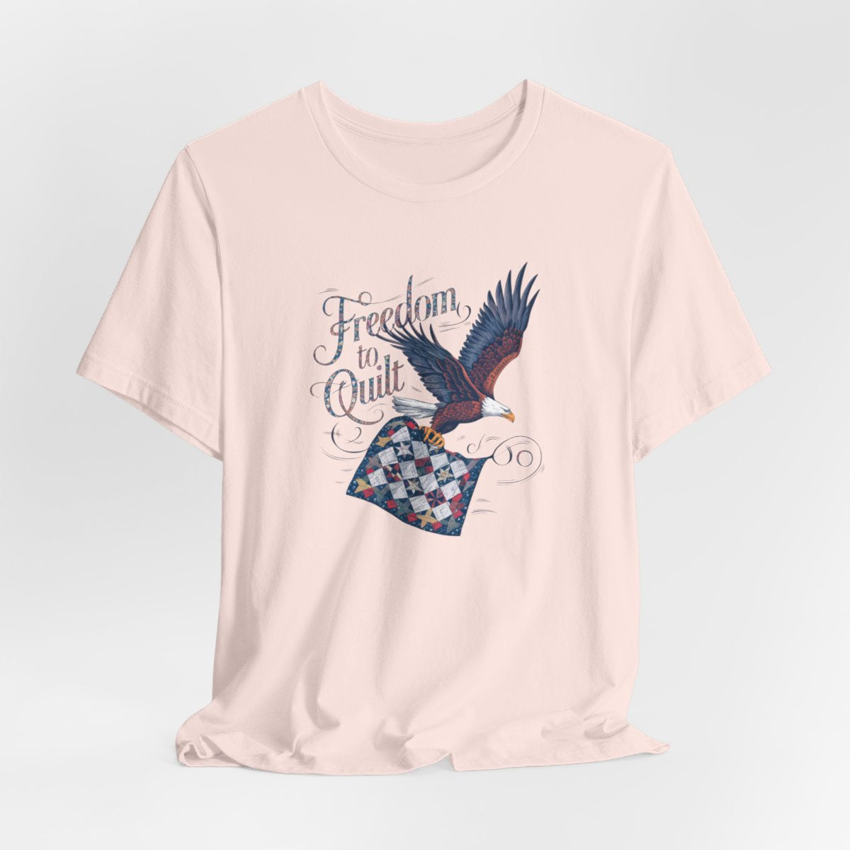 A Soft Pink T-shirt with detailed illustration of a bald eagle in flight, carrying a patchwork quilt. Text reads "Freedom to Quilt" in an ornate, patriotic font.