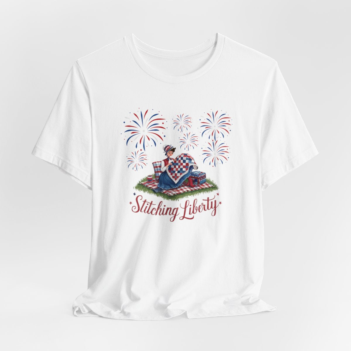 A White T-shirt with illustrated scene of a colonial woman quilting a patriotic blanket, surrounded by fireworks. Text reads "Stitching Liberty" in a decorative font.