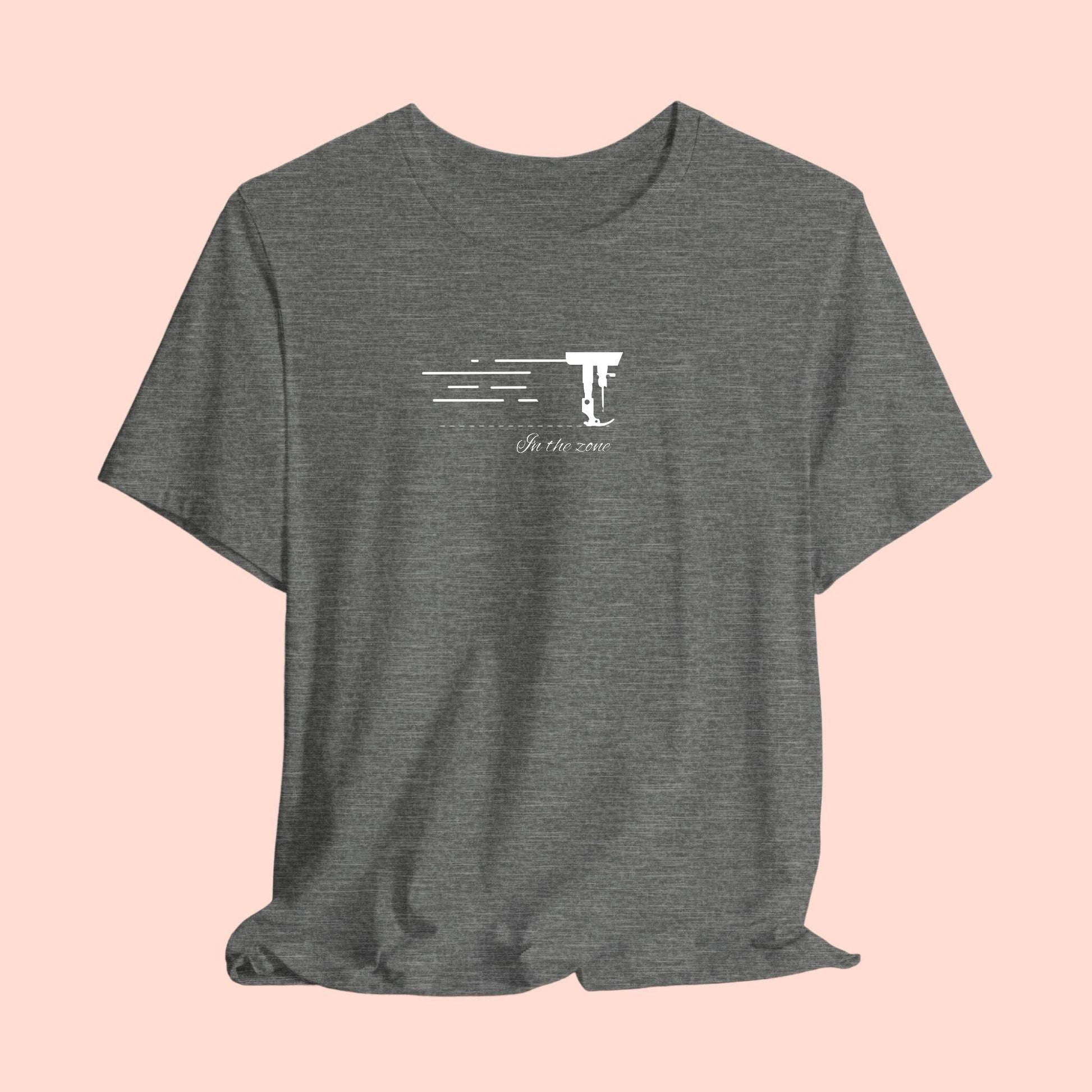 A T-shirt featuring a minimalist black silhouette of a sewing machine arm with motion lines. Below, the phrase "In the zone" is written in cursive script. The design conveys the focus and rhythm of quilting.