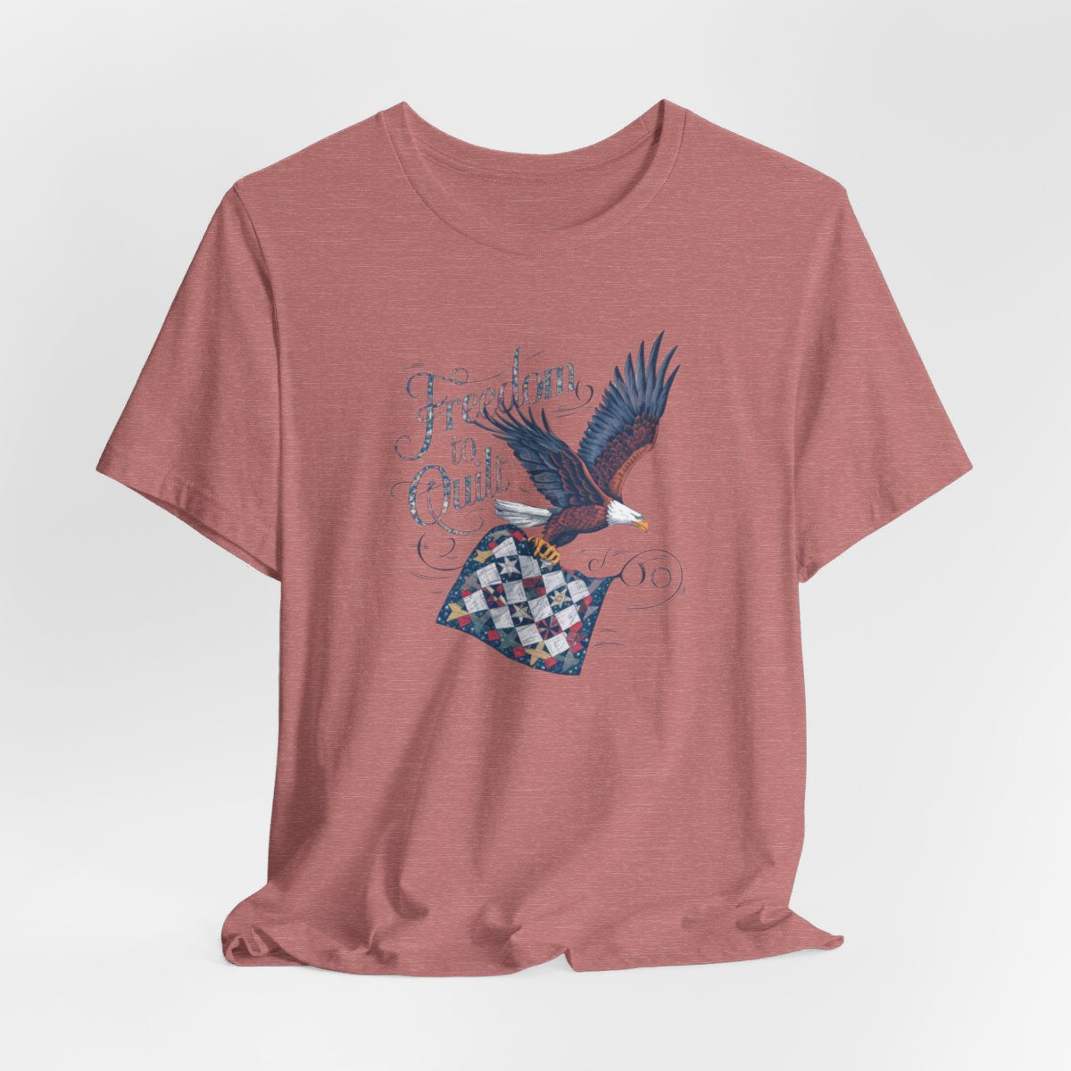 A Heather Mauve T-shirt with detailed illustration of a bald eagle in flight, carrying a patchwork quilt. Text reads "Freedom to Quilt" in an ornate, patriotic font.