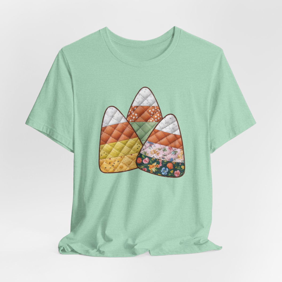 A Heather Mint Halloween Quilting T-Shirt featuring three large candy corn shapes made of quilted fabrics with various patterns. Each candy corn section features different quilting motifs in autumn colors. Playful take on Halloween candy and quilting craft.