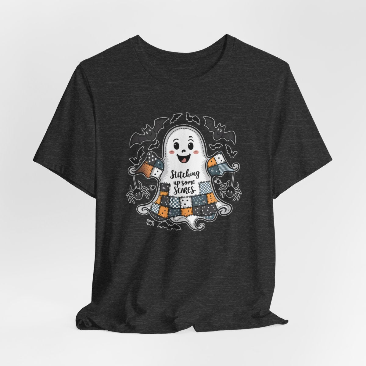 A Dark Grey Heather Halloween Quilting T-Shirt featuring a smiling ghost with quilted patchwork body. Halloween-themed quilt patterns. Cute bats and spiders around ghost. Text reads "Stitching up some Scares". Ideal for quilters who enjoy Halloween themes.