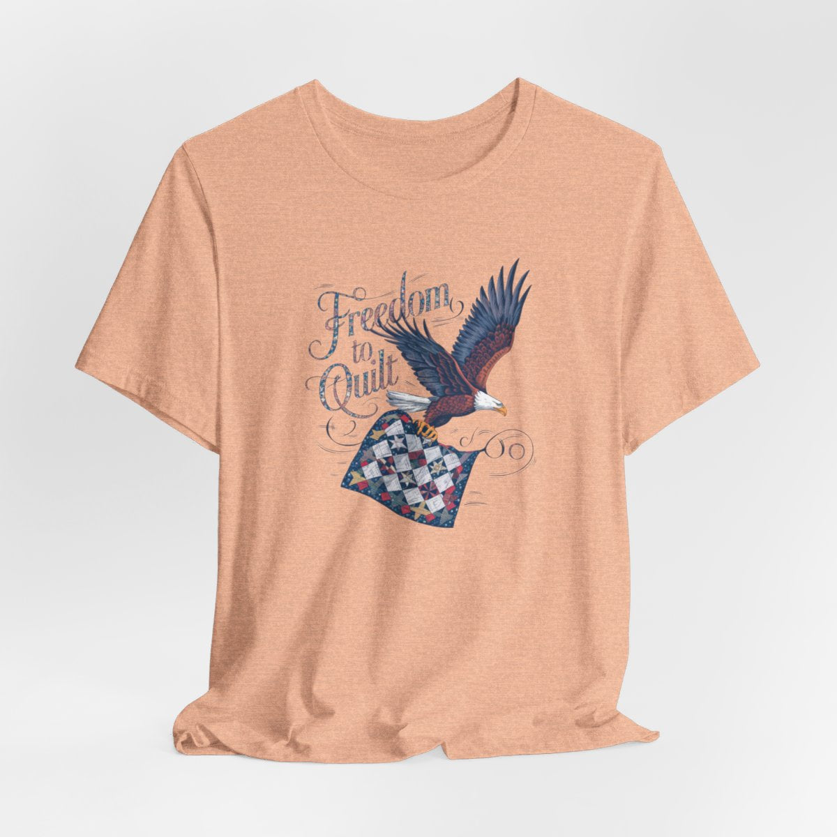 A Heather Peach T-shirt with detailed illustration of a bald eagle in flight, carrying a patchwork quilt. Text reads "Freedom to Quilt" in an ornate, patriotic font.