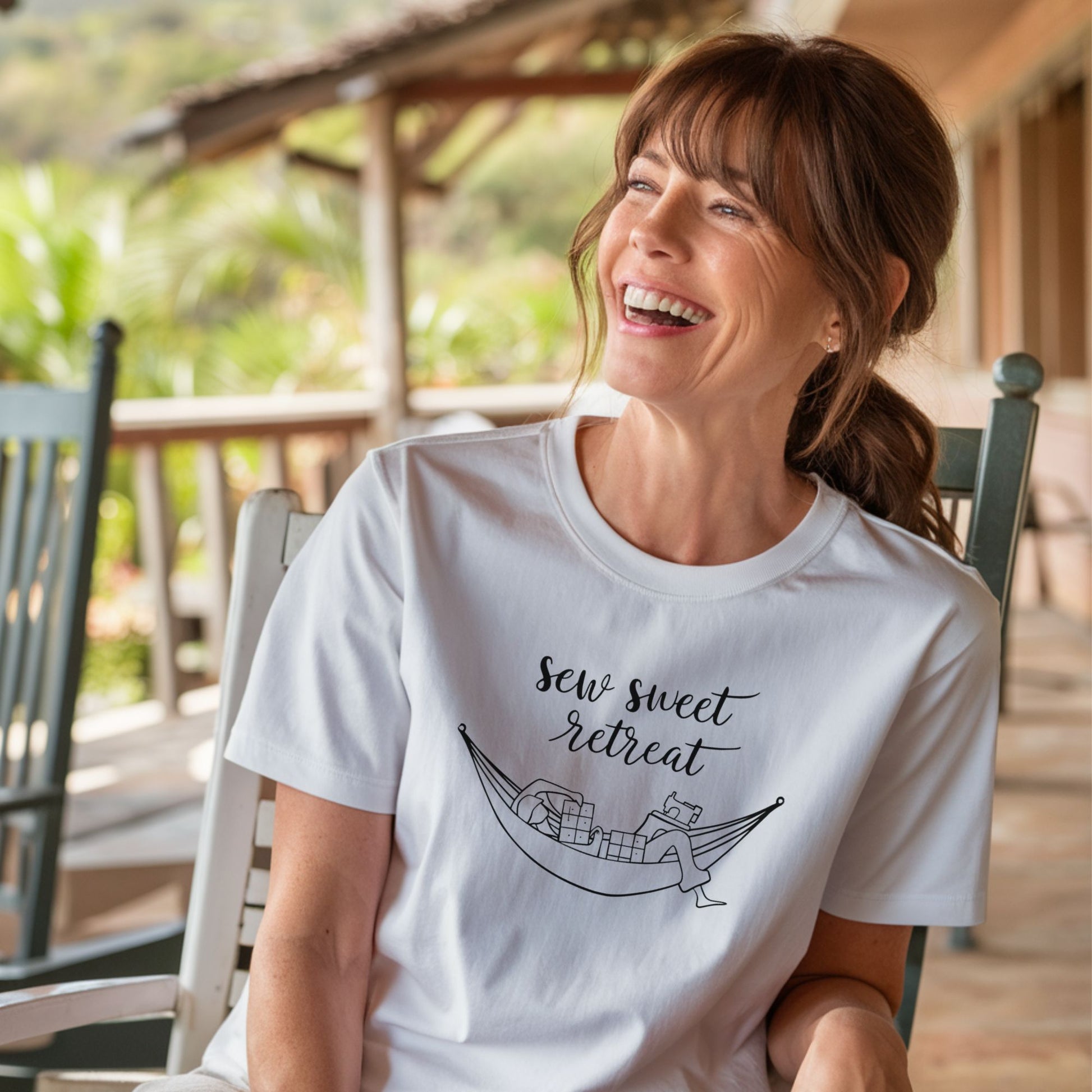 A T-Shirt with a black and white illustration of a hammock filled with quilting supplies like fabric squares and a sewing machine. Text above reads "Sew Sweet Retreat" in a flowing script font.