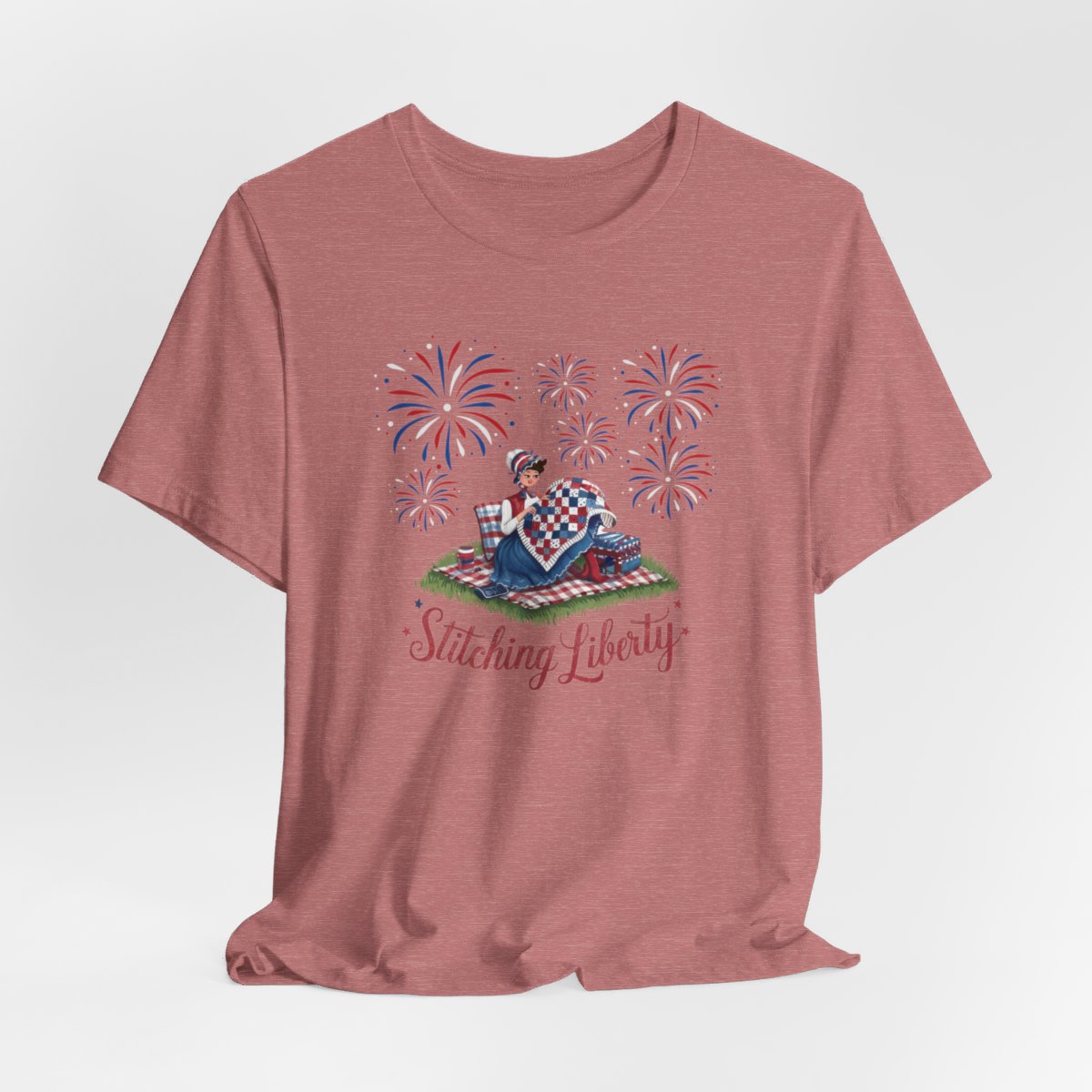 A Heather Mauve T-shirt with illustrated scene of a colonial woman quilting a patriotic blanket, surrounded by fireworks. Text reads "Stitching Liberty" in a decorative font.