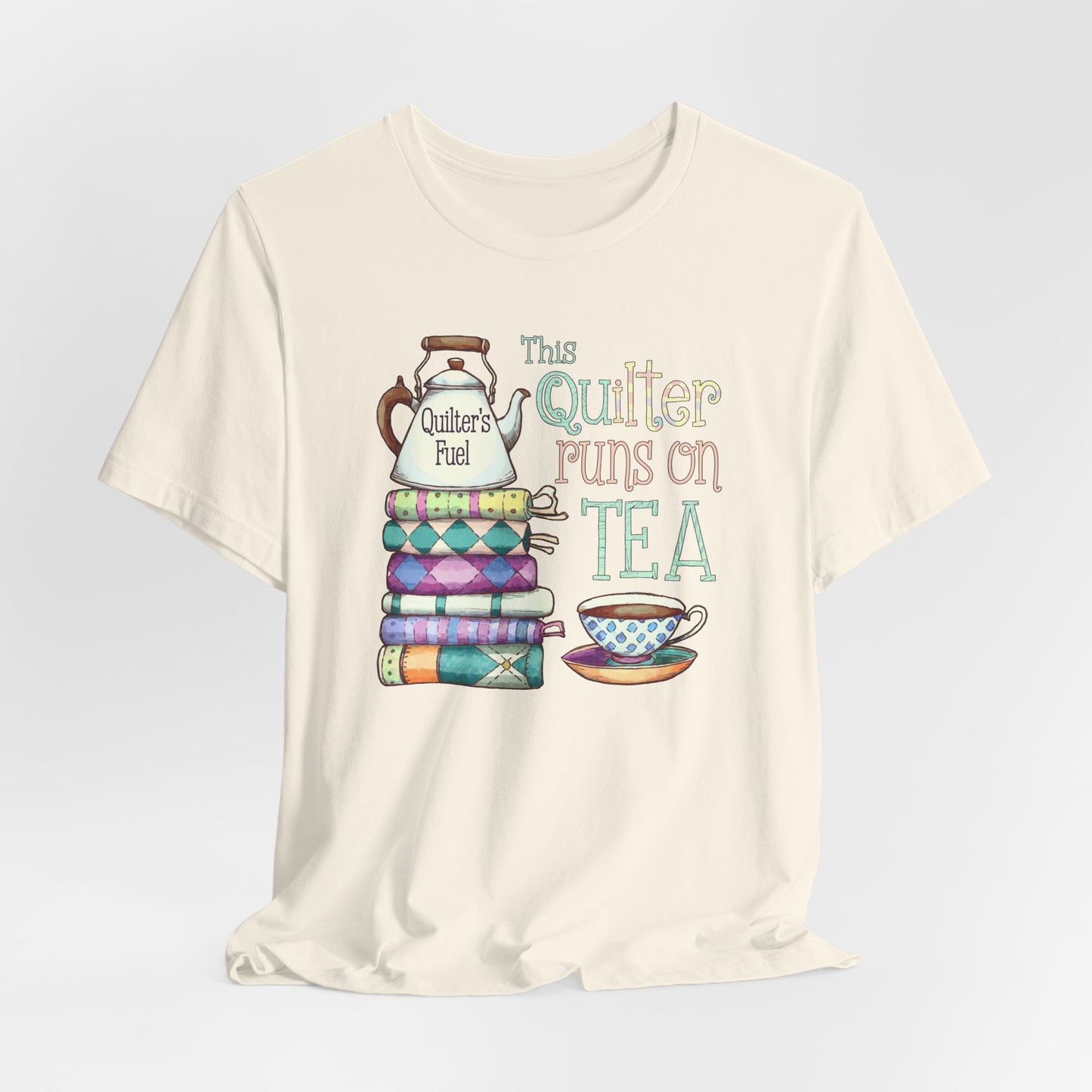 A Natural t-shirt featuring a whimsical design of a stack of patchwork quilts with a teapot labeled 'Quilter's Fuel' and the phrase 'This Quilter Runs on Tea,' ideal for tea-loving quilters.