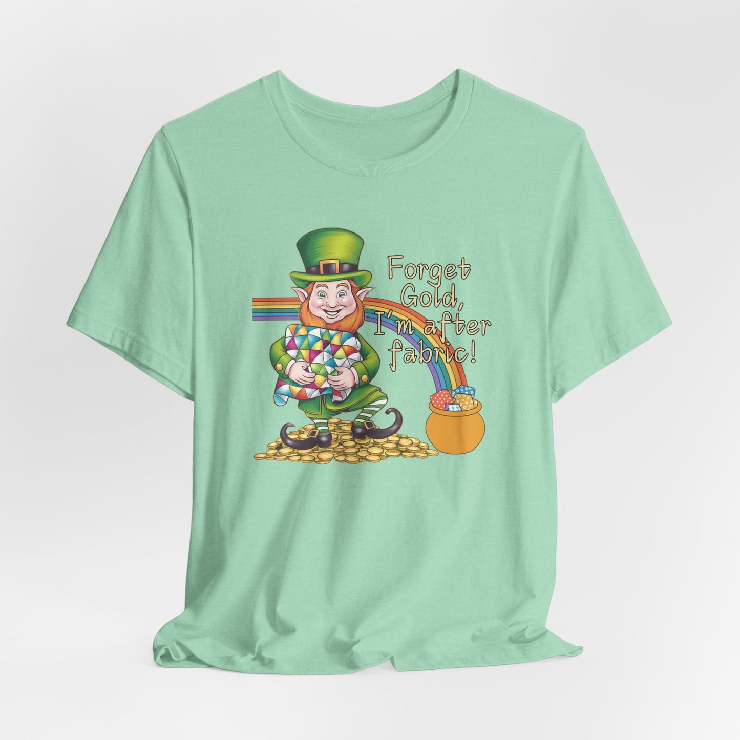 A Heather Mint short-sleeve t-shirt featuring a cheerful leprechaun holding a quilt, sitting on gold coins, with a rainbow leading to a fabric-filled pot and the phrase 'Forget Gold, I’m After Fabric!' Perfect for quilters celebrating St. Patrick’s Day.