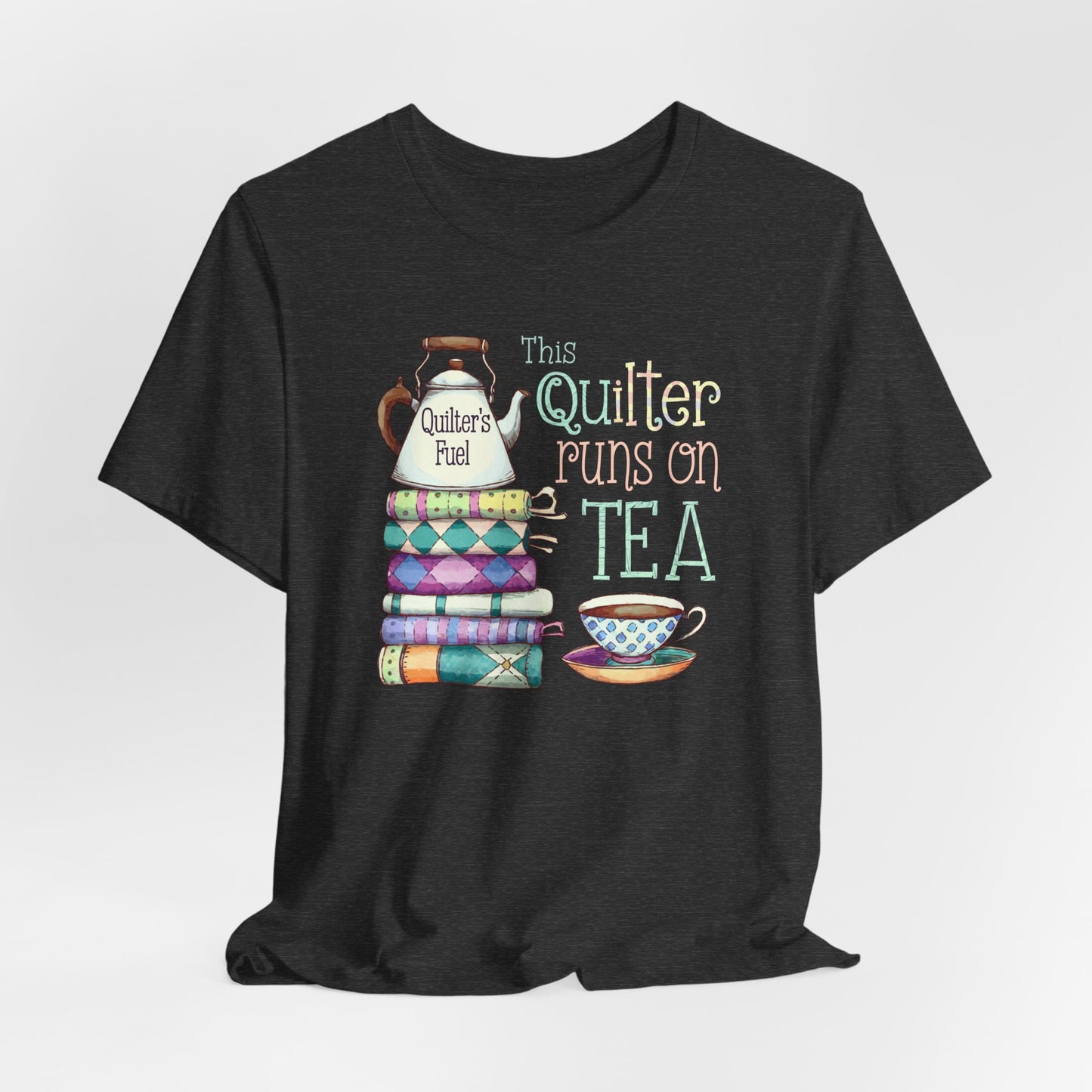 A Dark Grey Heather t-shirt featuring a whimsical design of a stack of patchwork quilts with a teapot labeled 'Quilter's Fuel' and the phrase 'This Quilter Runs on Tea,' ideal for tea-loving quilters.