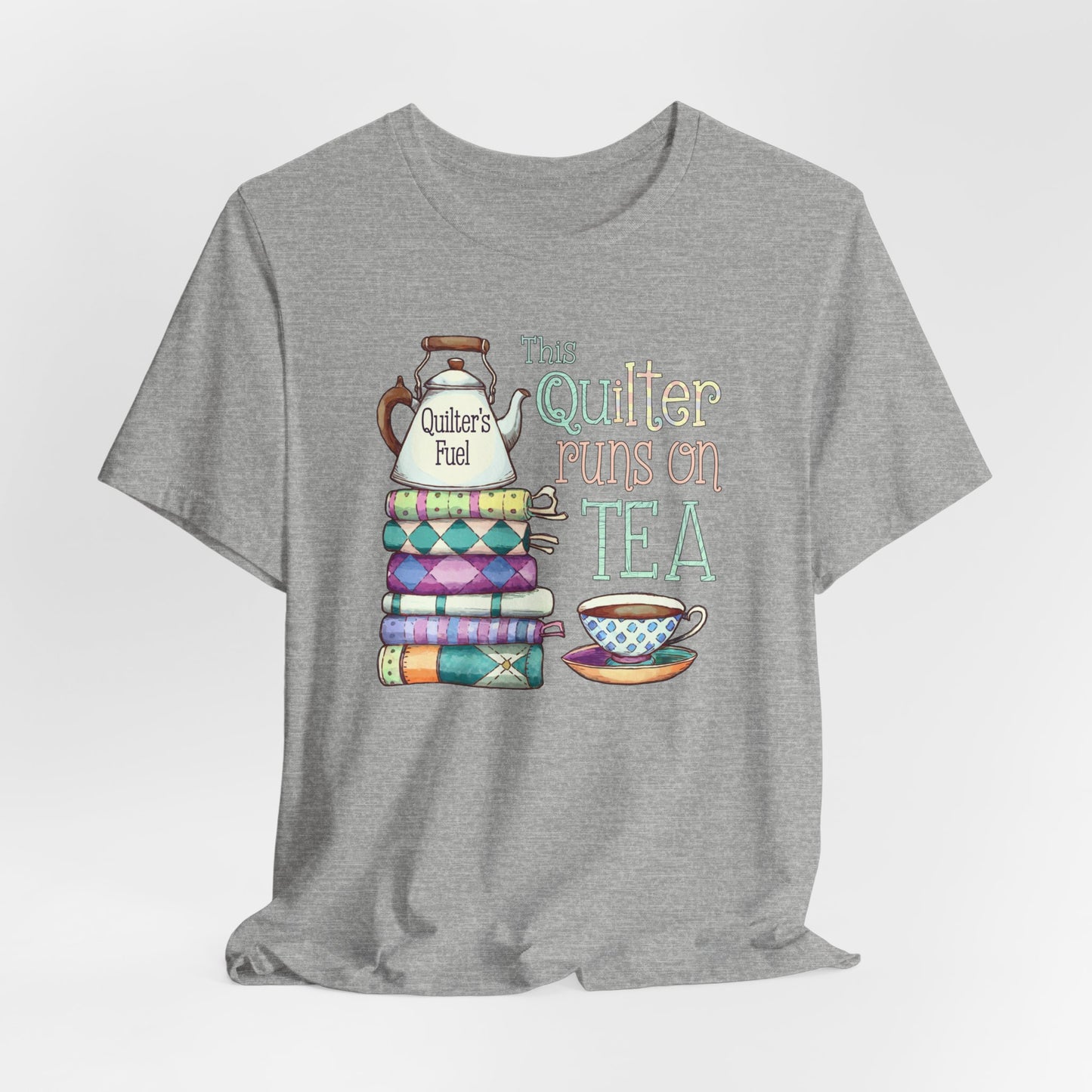 An Athletic Heather t-shirt featuring a whimsical design of a stack of patchwork quilts with a teapot labeled 'Quilter's Fuel' and the phrase 'This Quilter Runs on Tea,' ideal for tea-loving quilters.