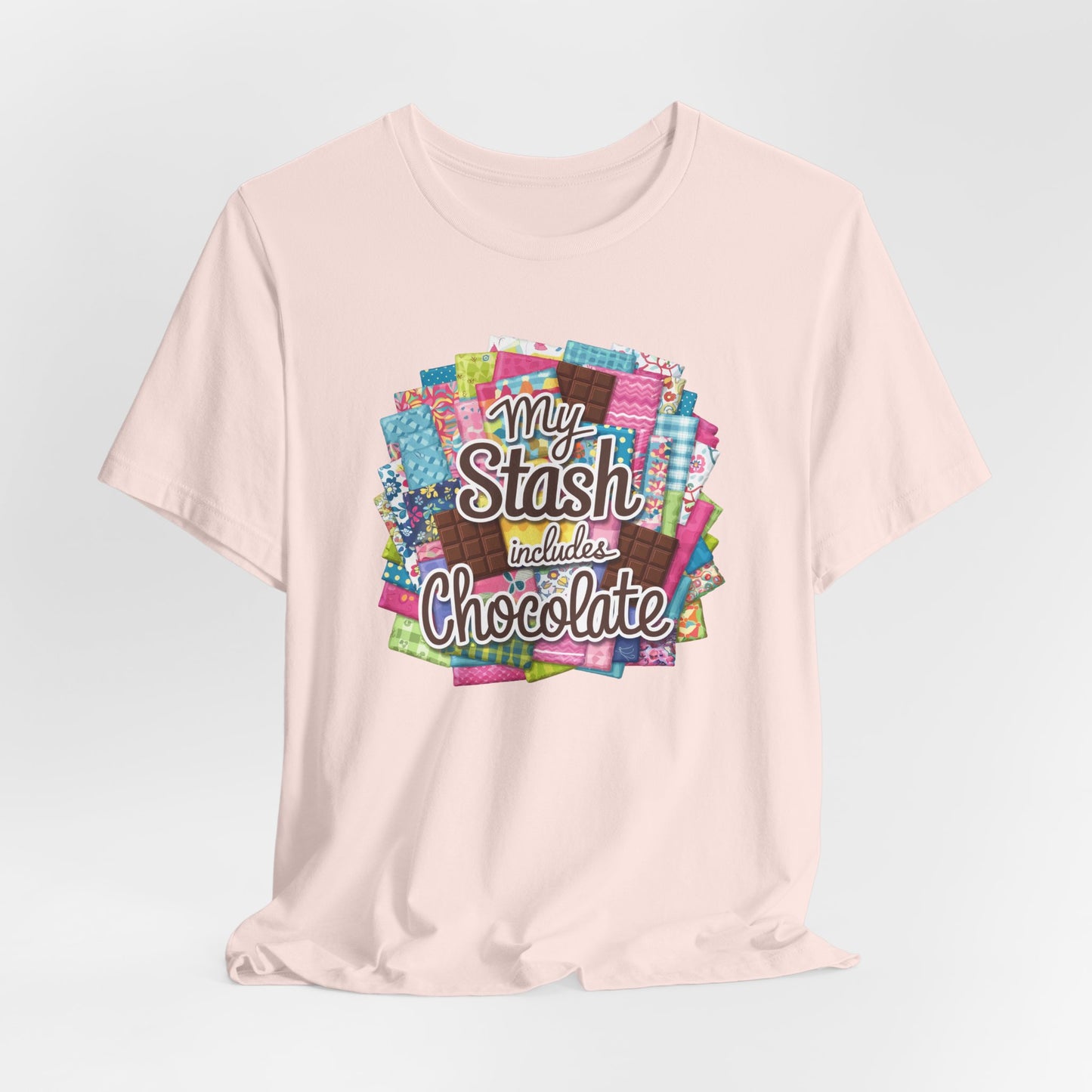 A Soft Pink t-shirt featuring a colorful design of a fabric stash mixed with chocolate bars and the phrase 'My Stash Includes Chocolate,' perfect for quilters and chocolate lovers