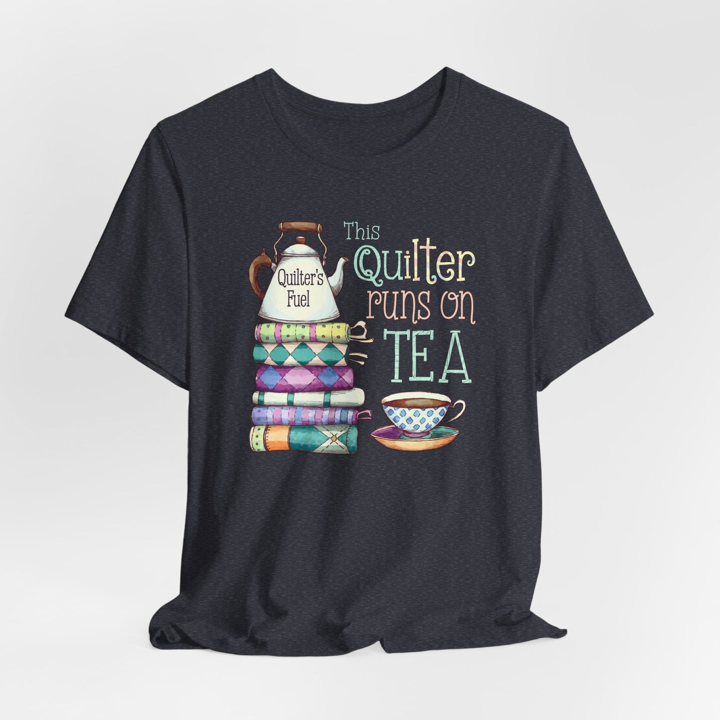 A Heather Navy t-shirt featuring a whimsical design of a stack of patchwork quilts with a teapot labeled 'Quilter's Fuel' and the phrase 'This Quilter Runs on Tea,' ideal for tea-loving quilters.