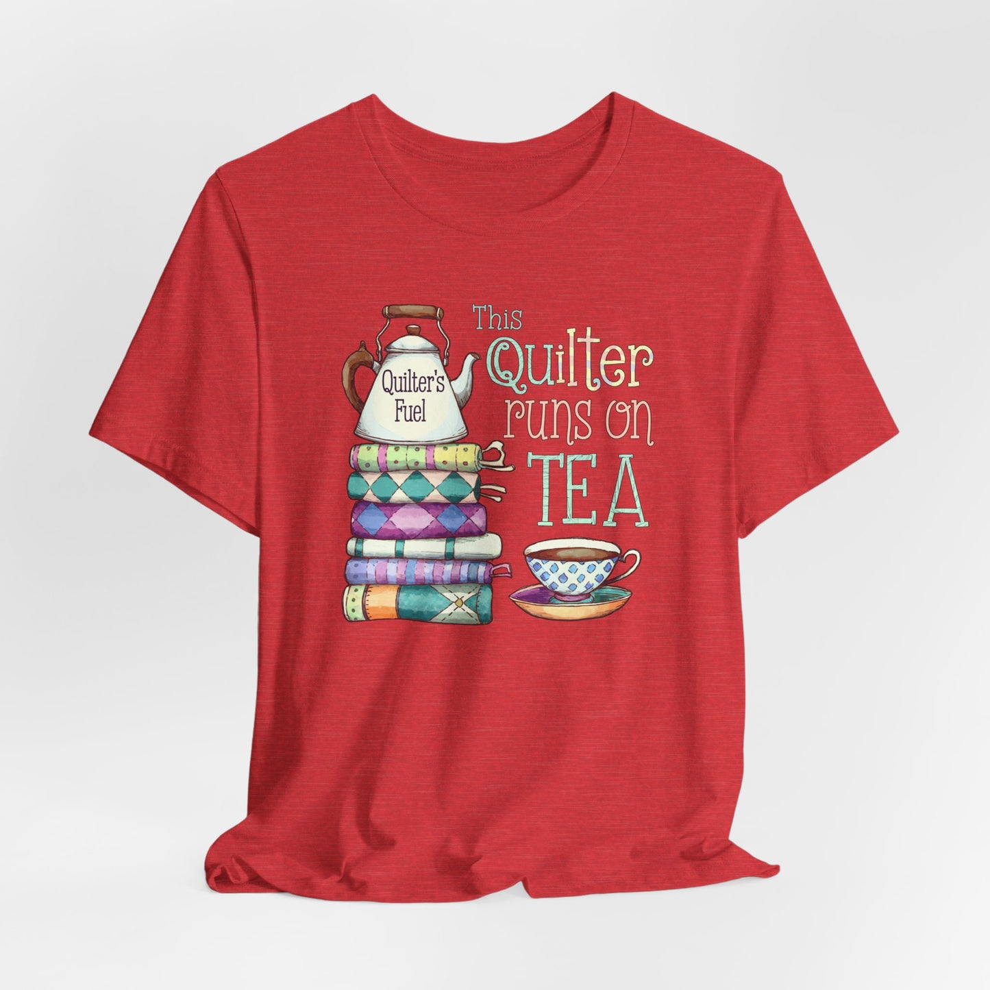 A Heather Red t-shirt featuring a whimsical design of a stack of patchwork quilts with a teapot labeled 'Quilter's Fuel' and the phrase 'This Quilter Runs on Tea,' ideal for tea-loving quilters.