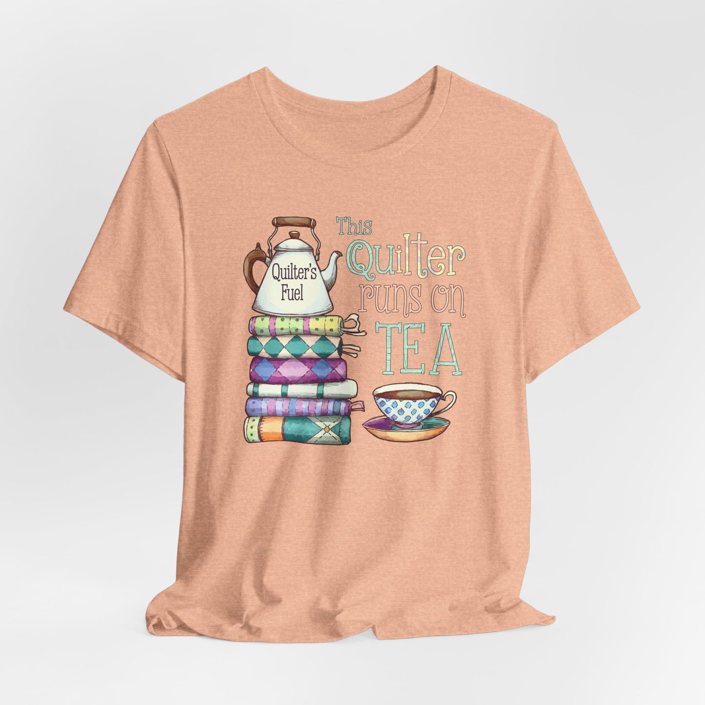 A Heather Peach t-shirt featuring a whimsical design of a stack of patchwork quilts with a teapot labeled 'Quilter's Fuel' and the phrase 'This Quilter Runs on Tea,' ideal for tea-loving quilters.