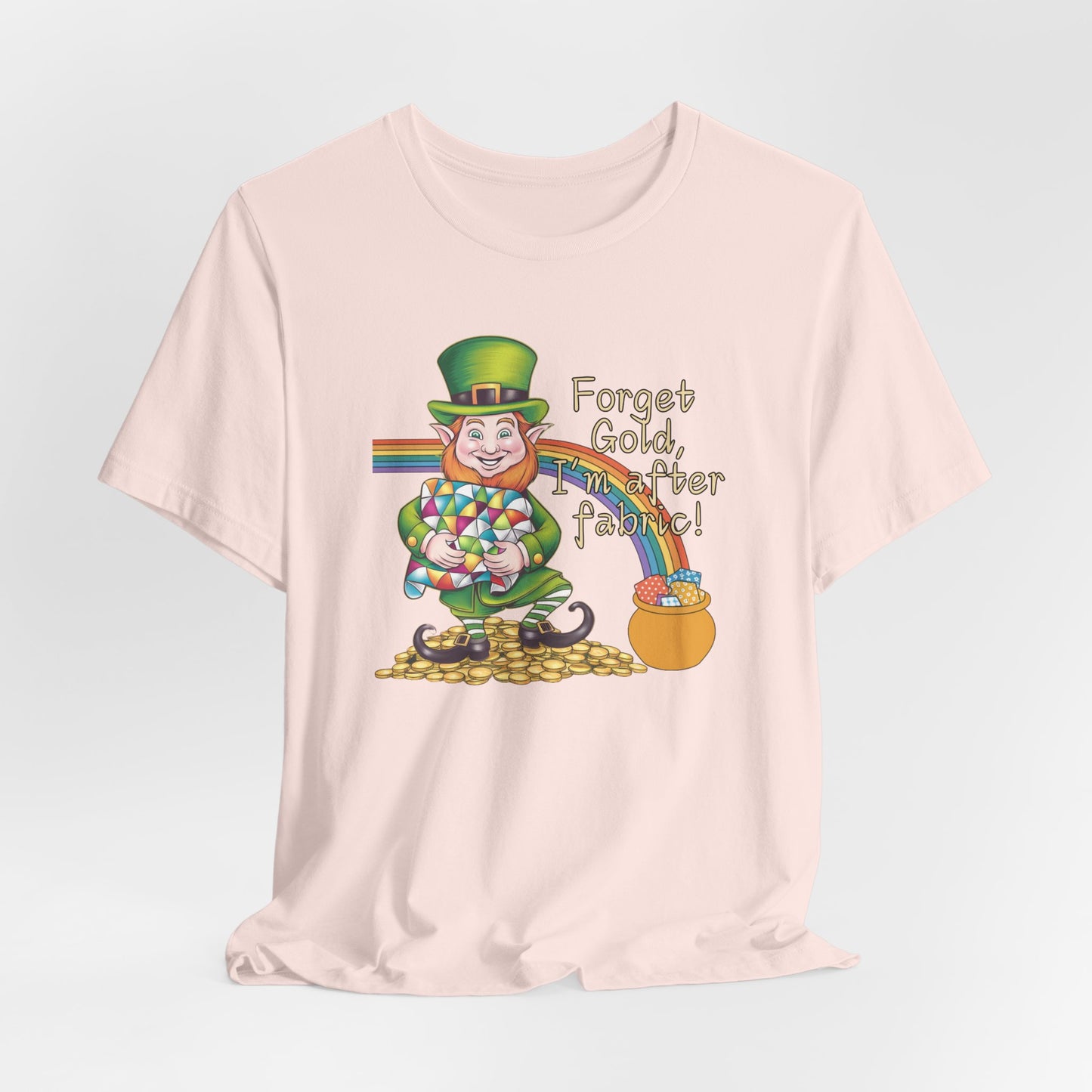 A Soft Pink short-sleeve t-shirt featuring a cheerful leprechaun holding a quilt, sitting on gold coins, with a rainbow leading to a fabric-filled pot and the phrase 'Forget Gold, I’m After Fabric!' Perfect for quilters celebrating St. Patrick’s Day.