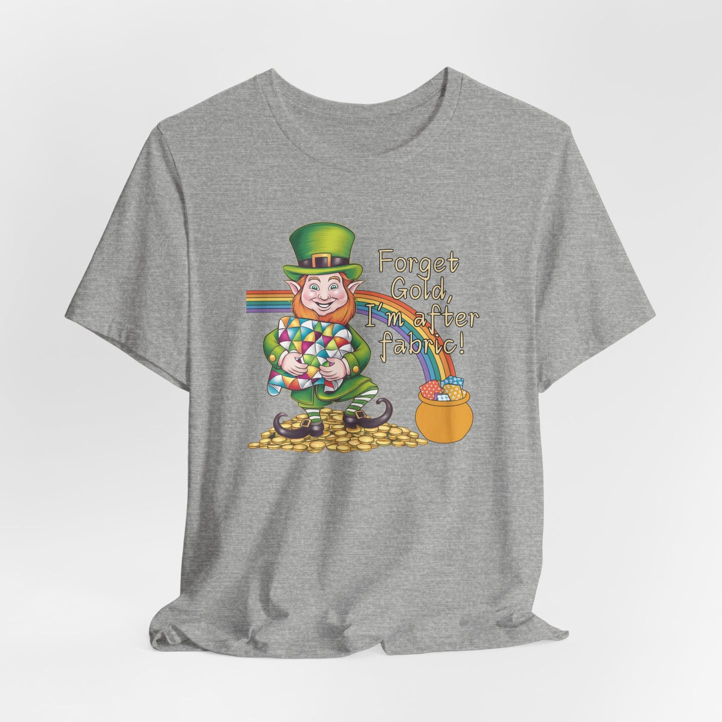 An Athletic Heather short-sleeve t-shirt featuring a cheerful leprechaun holding a quilt, sitting on gold coins, with a rainbow leading to a fabric-filled pot and the phrase 'Forget Gold, I’m After Fabric!' Perfect for quilters celebrating St. Patrick’s Day.