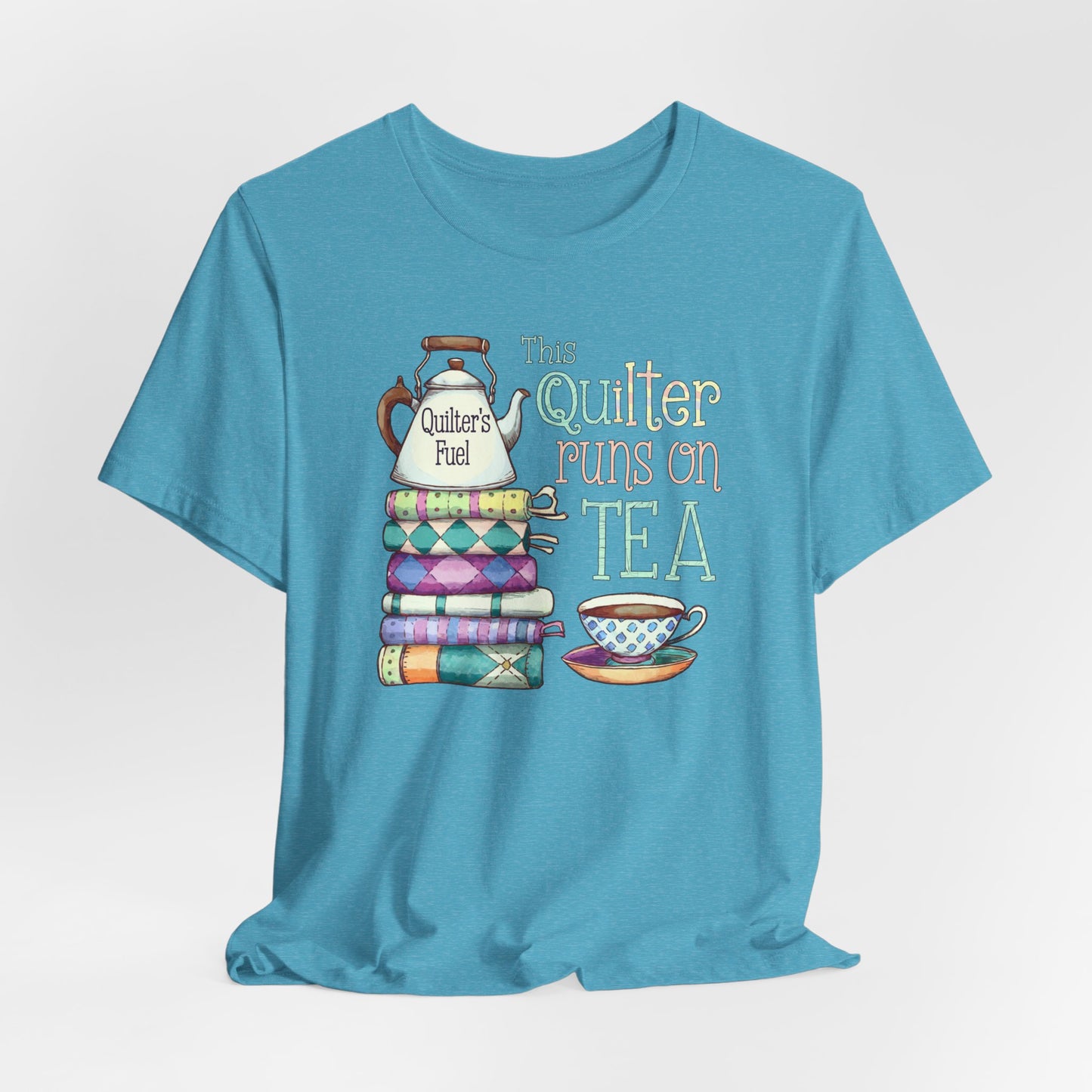 A Heather Aqua t-shirt featuring a whimsical design of a stack of patchwork quilts with a teapot labeled 'Quilter's Fuel' and the phrase 'This Quilter Runs on Tea,' ideal for tea-loving quilters.