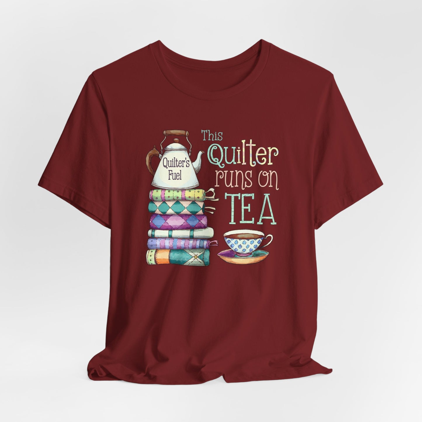 A Cardinal t-shirt featuring a whimsical design of a stack of patchwork quilts with a teapot labeled 'Quilter's Fuel' and the phrase 'This Quilter Runs on Tea,' ideal for tea-loving quilters.