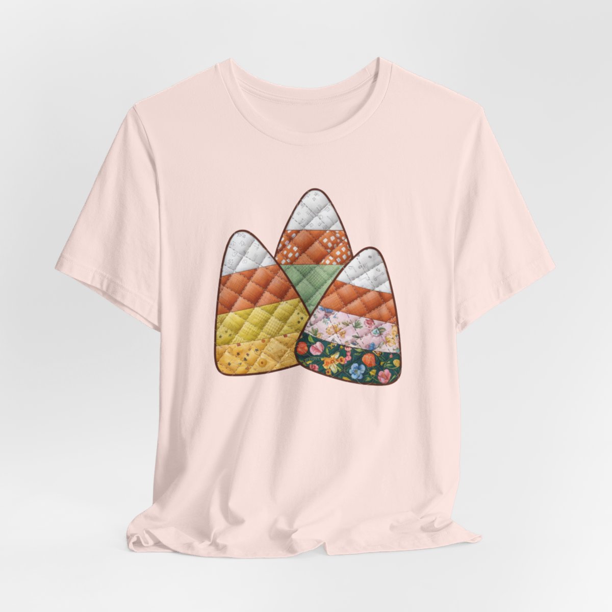 A Soft Pink Halloween Quilting T-Shirt featuring three large candy corn shapes made of quilted fabrics with various patterns. Each candy corn section features different quilting motifs in autumn colors. Playful take on Halloween candy and quilting craft.