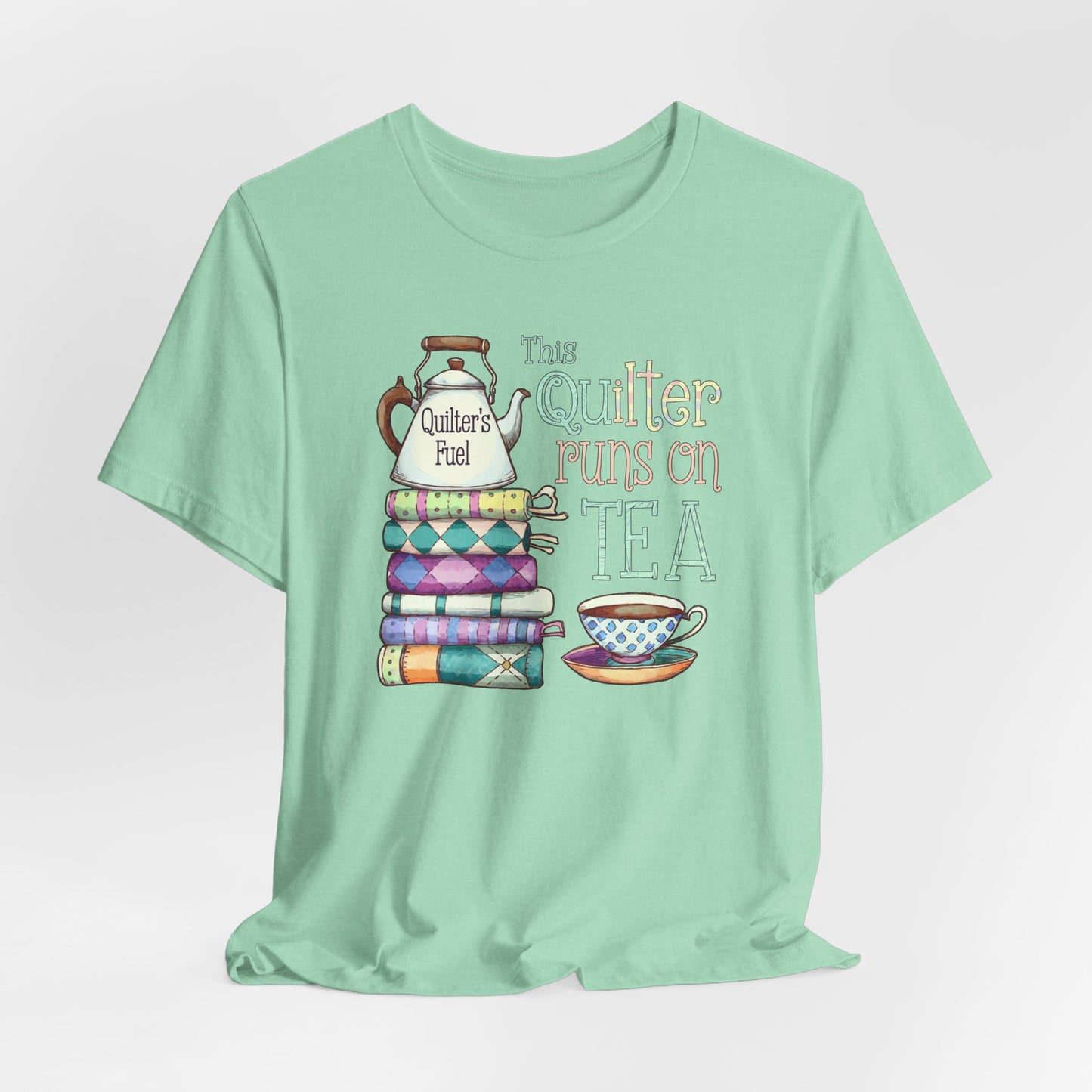 A Heather Mint t-shirt featuring a whimsical design of a stack of patchwork quilts with a teapot labeled 'Quilter's Fuel' and the phrase 'This Quilter Runs on Tea,' ideal for tea-loving quilters.