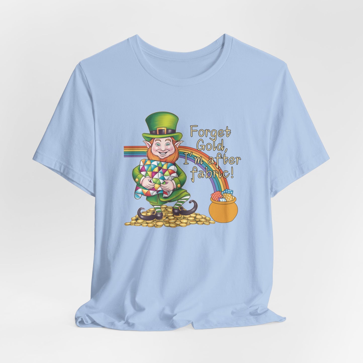 A Baby Blue short-sleeve t-shirt featuring a cheerful leprechaun holding a quilt, sitting on gold coins, with a rainbow leading to a fabric-filled pot and the phrase 'Forget Gold, I’m After Fabric!' Perfect for quilters celebrating St. Patrick’s Day.