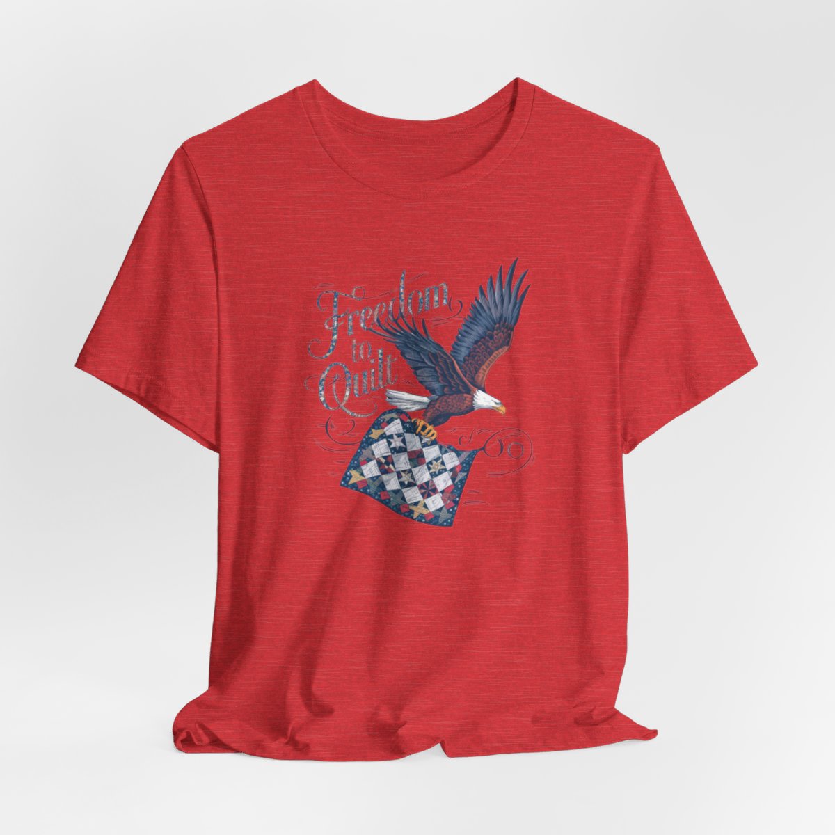 A Heather Red T-shirt with detailed illustration of a bald eagle in flight, carrying a patchwork quilt. Text reads "Freedom to Quilt" in an ornate, patriotic font.