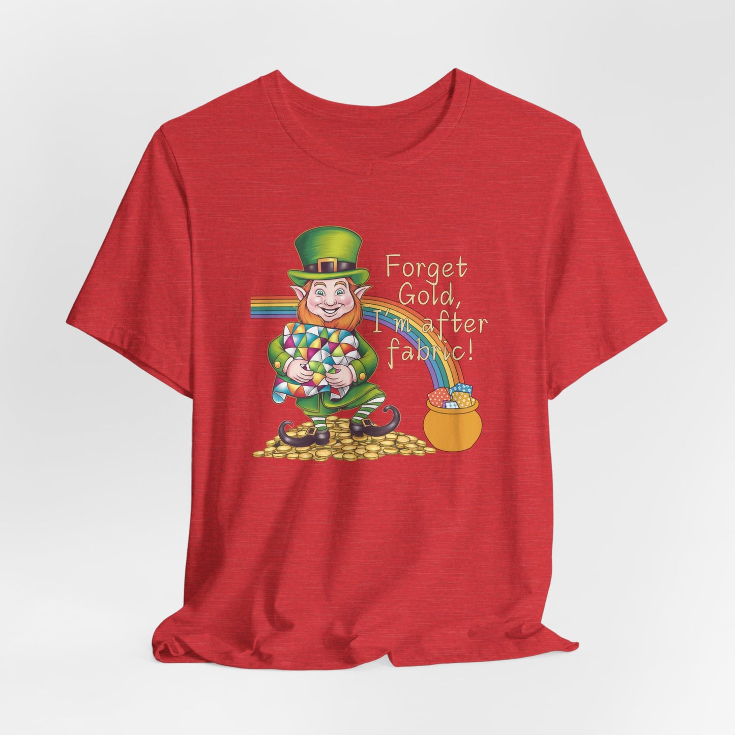 A Heather Red short-sleeve t-shirt featuring a cheerful leprechaun holding a quilt, sitting on gold coins, with a rainbow leading to a fabric-filled pot and the phrase 'Forget Gold, I’m After Fabric!' Perfect for quilters celebrating St. Patrick’s Day.