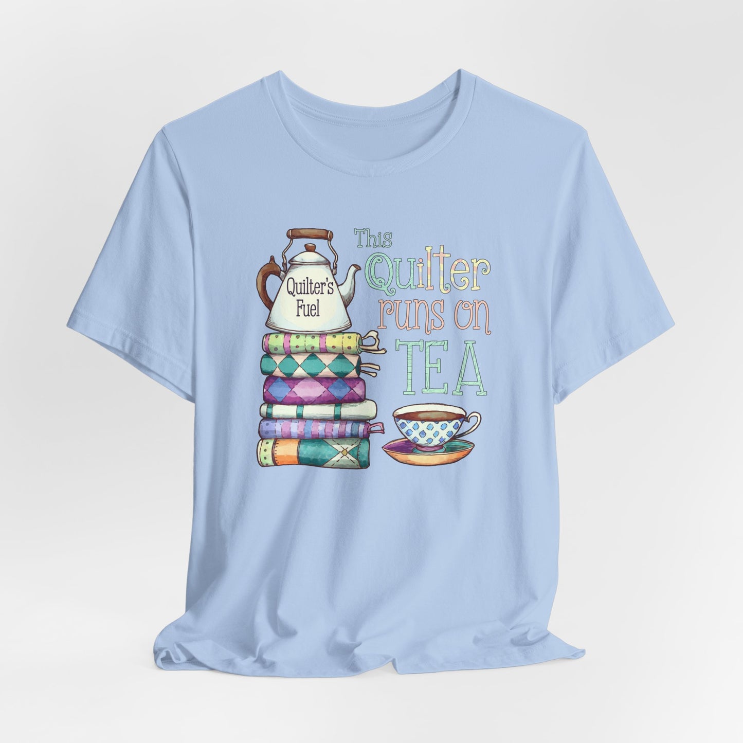 A Baby Blue t-shirt featuring a whimsical design of a stack of patchwork quilts with a teapot labeled 'Quilter's Fuel' and the phrase 'This Quilter Runs on Tea,' ideal for tea-loving quilters.