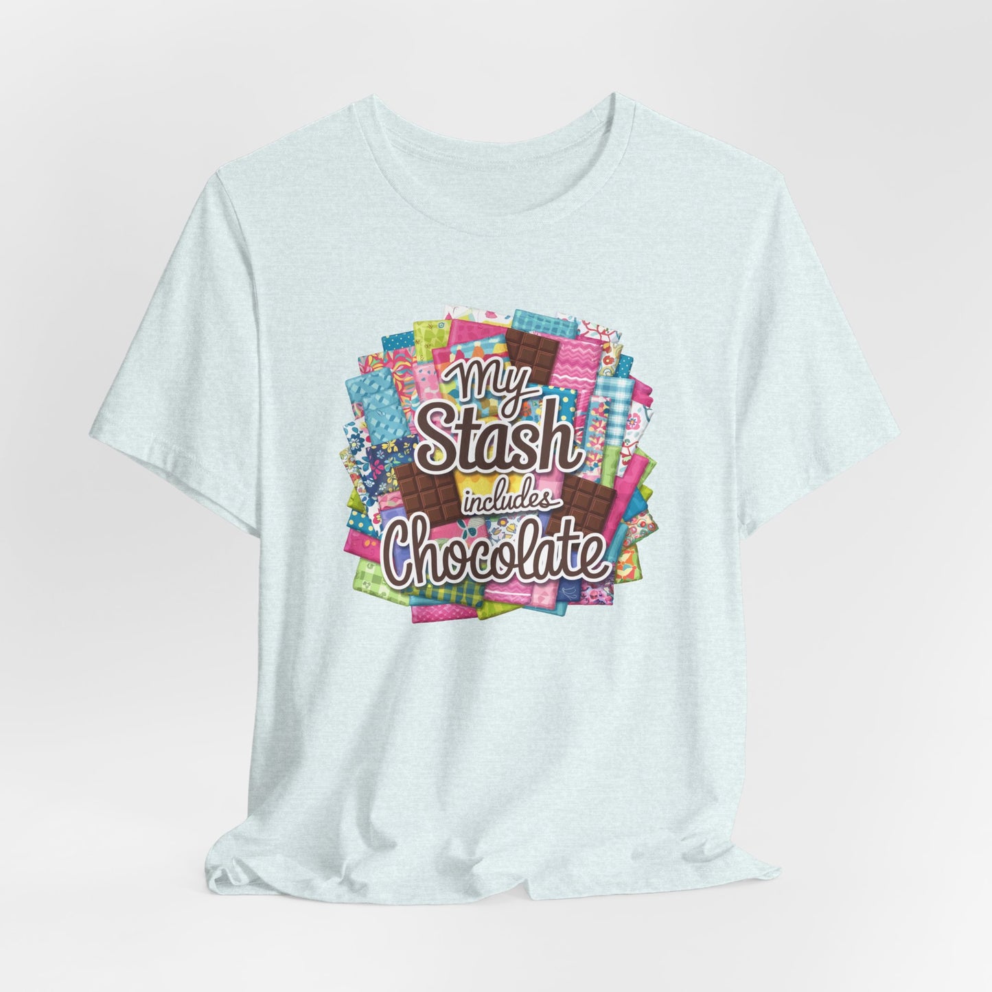 A Heather Ice Blue t-shirt featuring a colorful design of a fabric stash mixed with chocolate bars and the phrase 'My Stash Includes Chocolate,' perfect for quilters and chocolate lovers