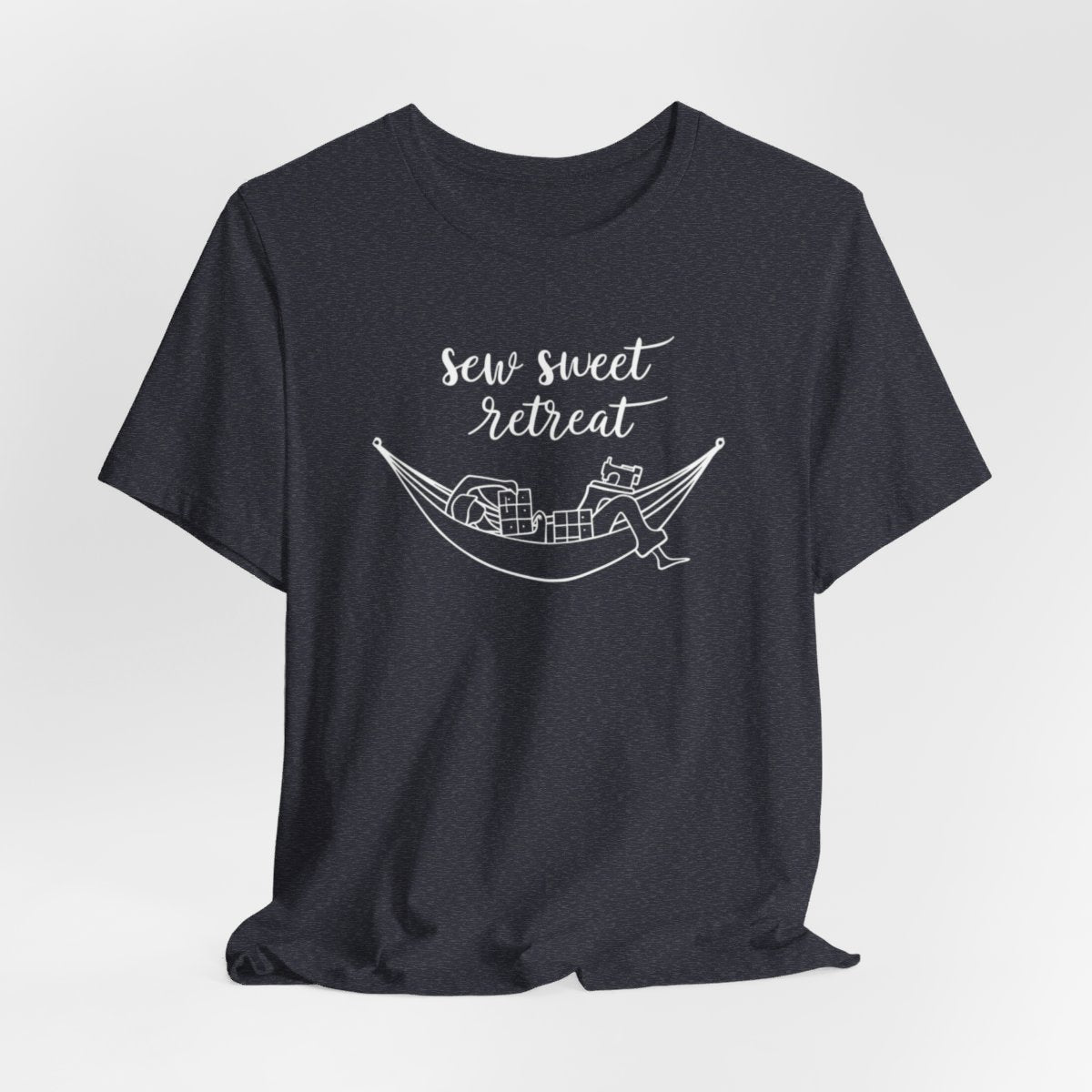 A Heather Navy T-Shirt with a black and white illustration of a hammock filled with quilting supplies like fabric squares and a sewing machine. Text above reads "Sew Sweet Retreat" in a flowing script font.