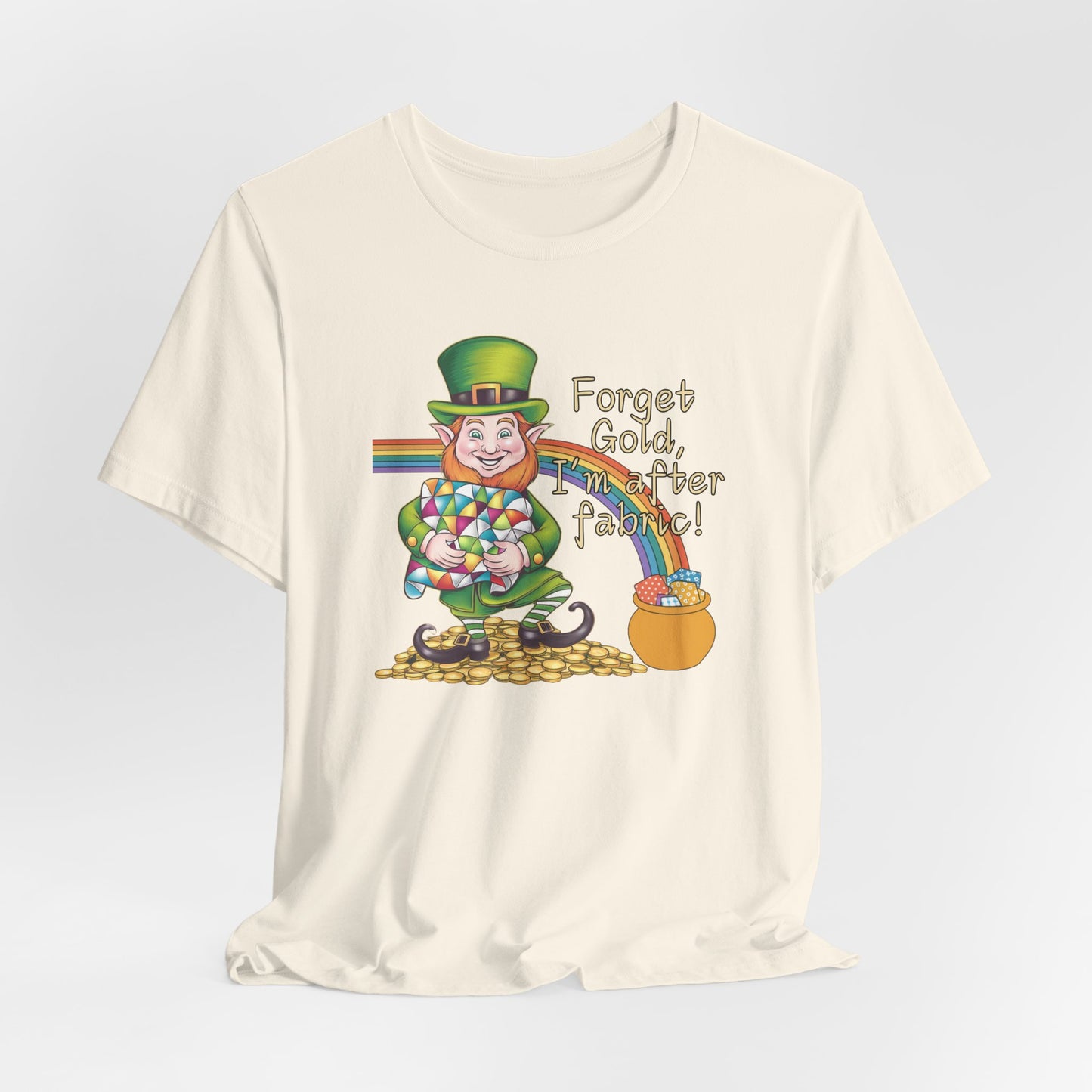 A Natural short-sleeve t-shirt featuring a cheerful leprechaun holding a quilt, sitting on gold coins, with a rainbow leading to a fabric-filled pot and the phrase 'Forget Gold, I’m After Fabric!' Perfect for quilters celebrating St. Patrick’s Day.