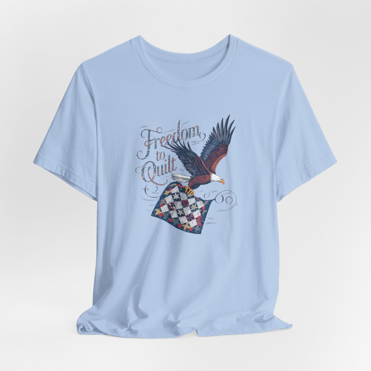 A Baby Blue T-shirt with detailed illustration of a bald eagle in flight, carrying a patchwork quilt. Text reads "Freedom to Quilt" in an ornate, patriotic font.