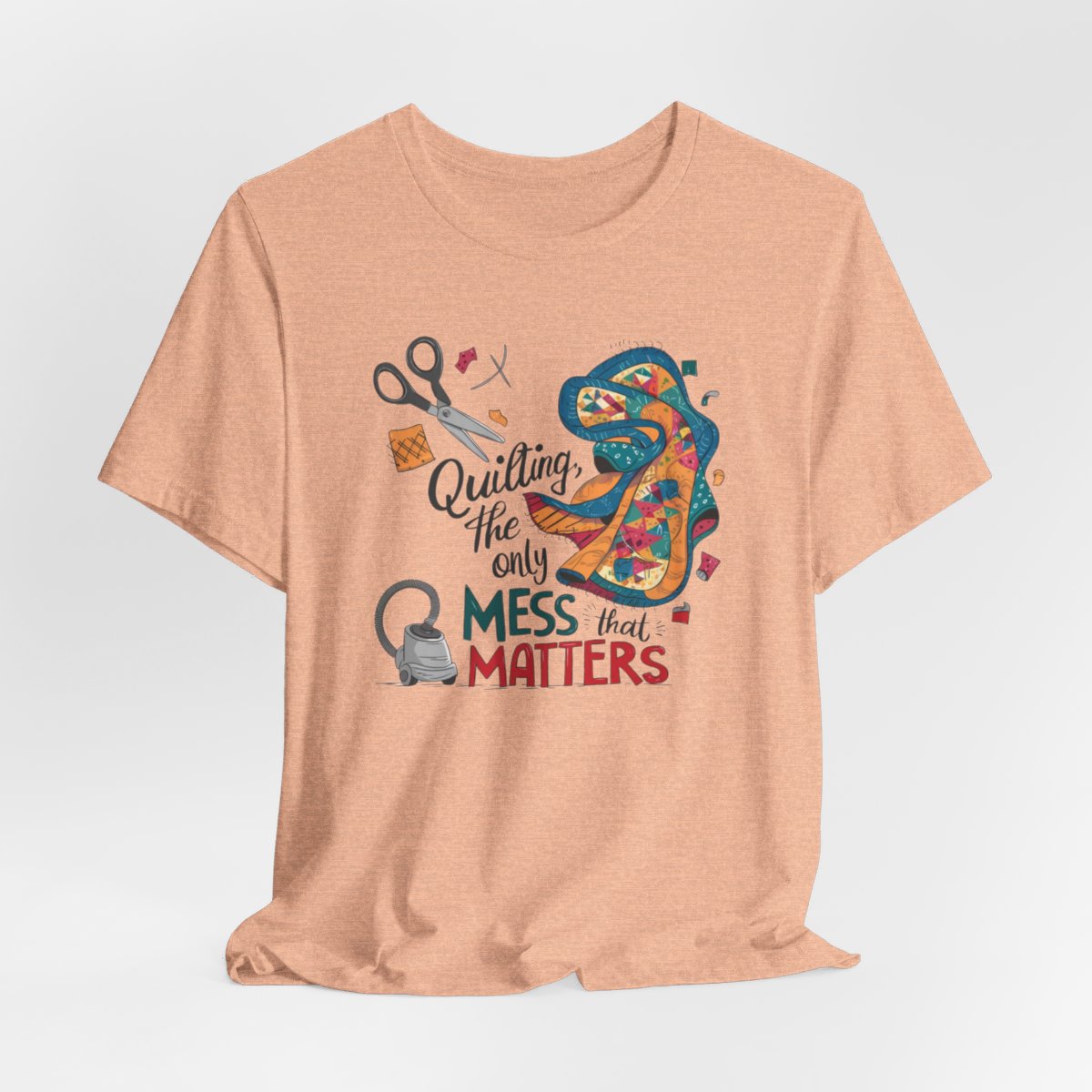 A Heather Peach fun T-Shirt with the phrase Quilting, the Only Mess that Matters and illustrations of fabric scraps, scissors, and a vacuum cleaner. Colorful quilt patterns swirl around the text, creating a playful and relatable design for quilting enthusiasts.