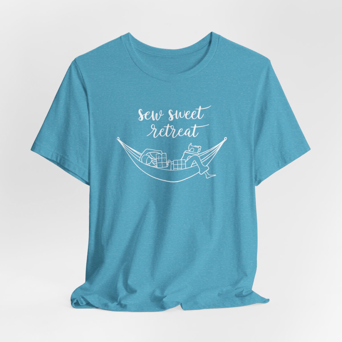 A Heather Aqua T-Shirt with a black and white illustration of a hammock filled with quilting supplies like fabric squares and a sewing machine. Text above reads "Sew Sweet Retreat" in a flowing script font.