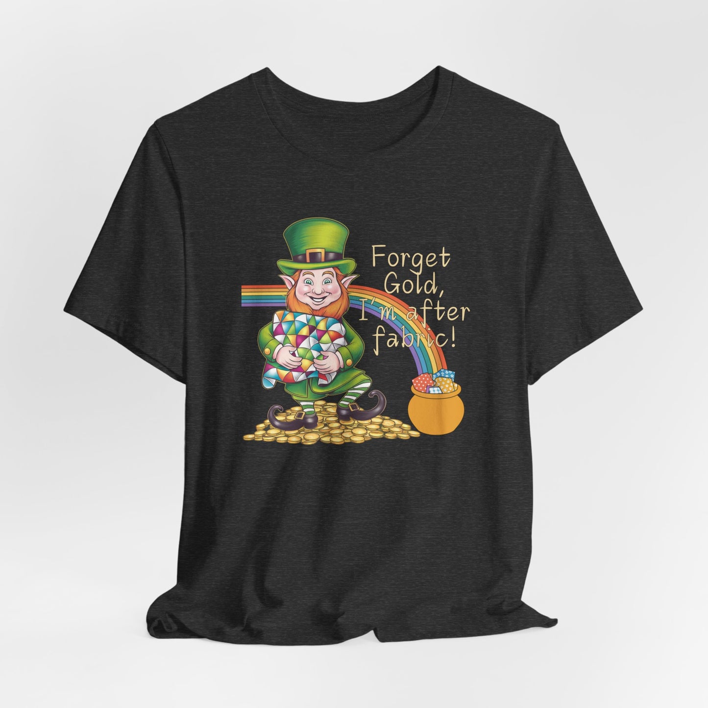 A Dark Grey Heather short-sleeve t-shirt featuring a cheerful leprechaun holding a quilt, sitting on gold coins, with a rainbow leading to a fabric-filled pot and the phrase 'Forget Gold, I’m After Fabric!' Perfect for quilters celebrating St. Patrick’s Day.