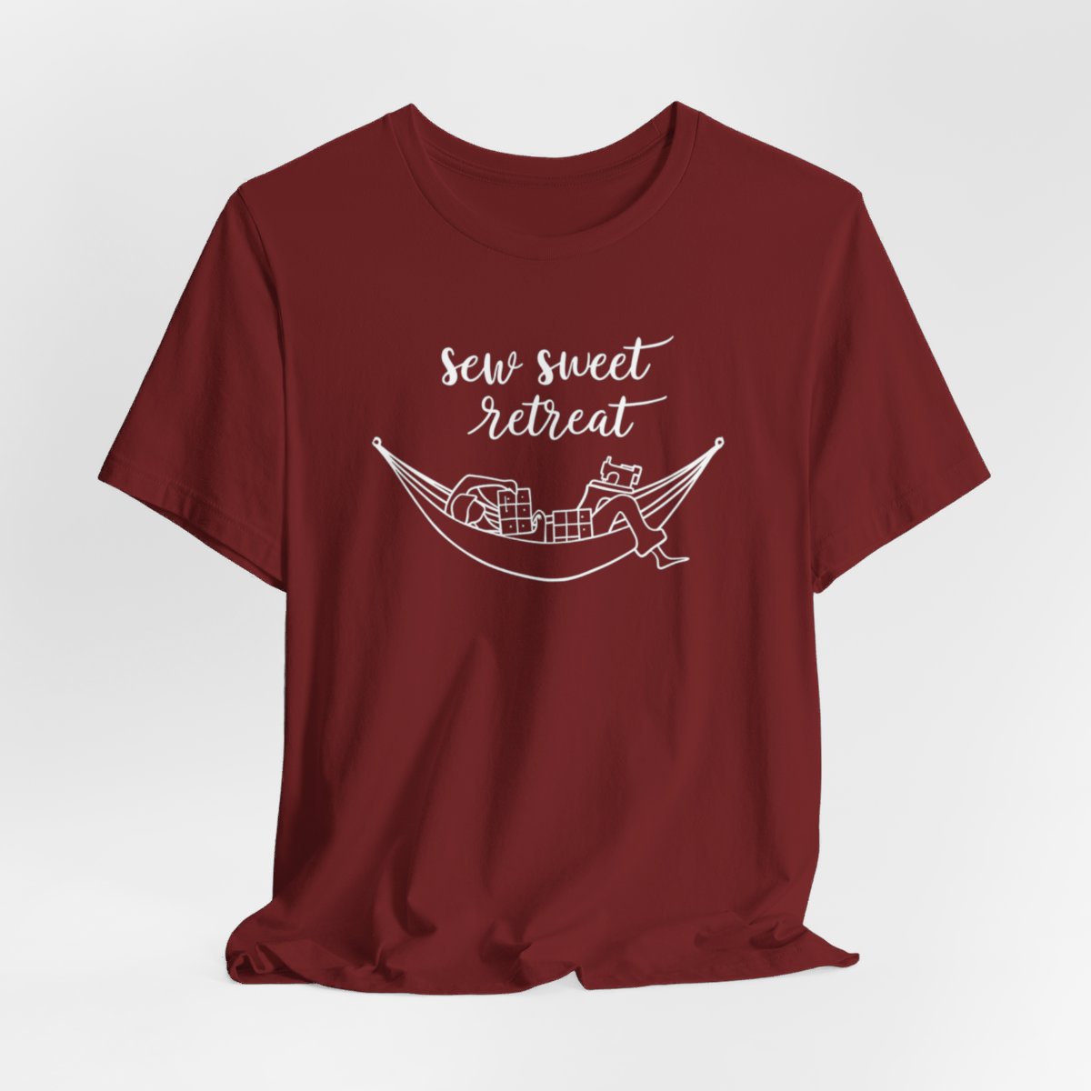 A Cardinal T-Shirt with a black and white illustration of a hammock filled with quilting supplies like fabric squares and a sewing machine. Text above reads "Sew Sweet Retreat" in a flowing script font.