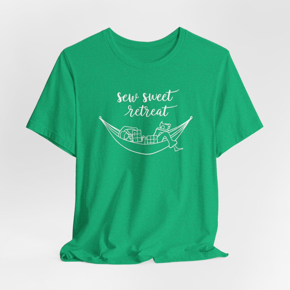 A Heather Kelly T-Shirt with a black and white illustration of a hammock filled with quilting supplies like fabric squares and a sewing machine. Text above reads "Sew Sweet Retreat" in a flowing script font.