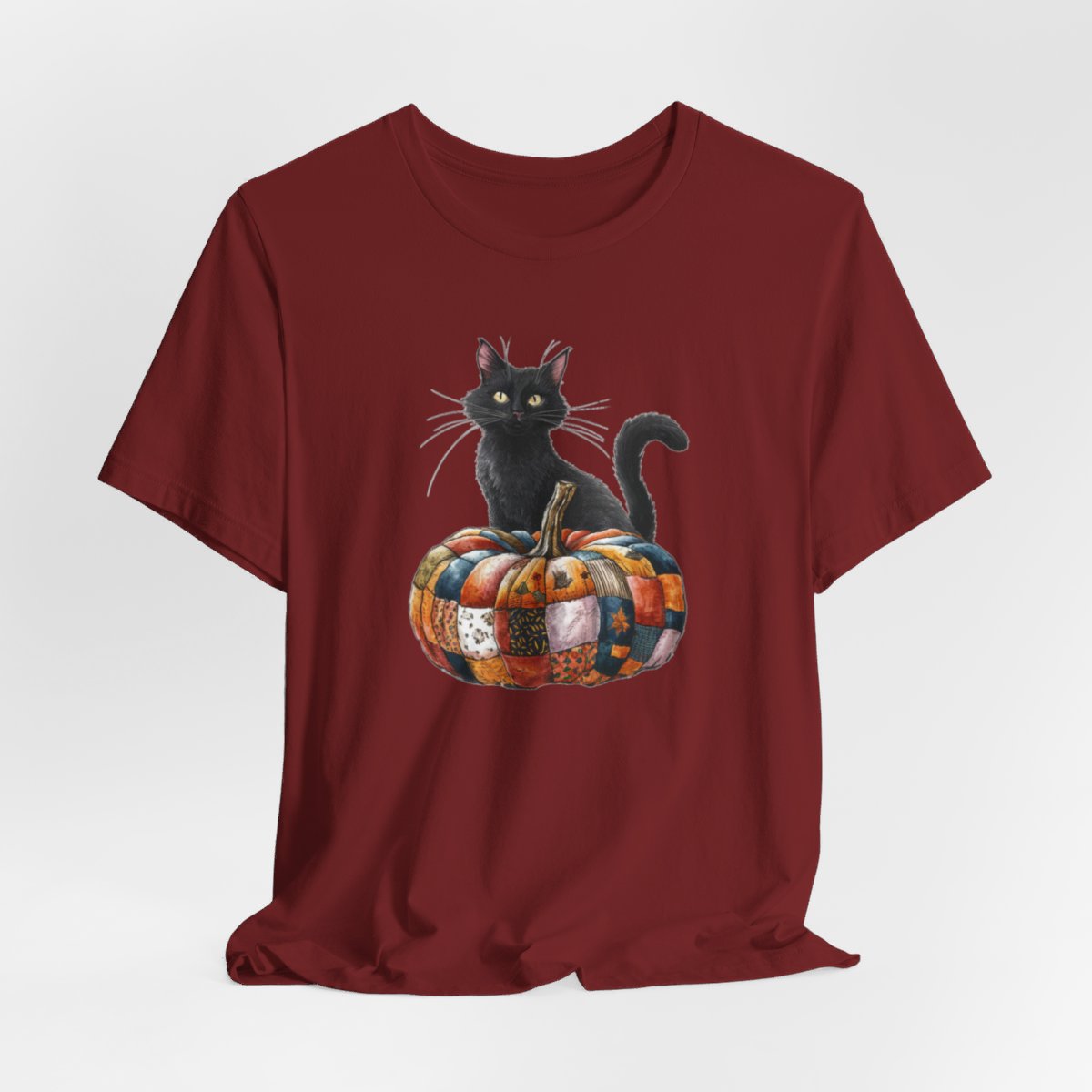 A Cardinal Halloween Quilting T-Shirt featuring a black cat sitting on colorful patchwork quilt pumpkin. Cat has yellow eyes and long whiskers. Pumpkin features various quilt patterns in autumn colors. Halloween-themed t-shirt design for quilting enthusiasts.