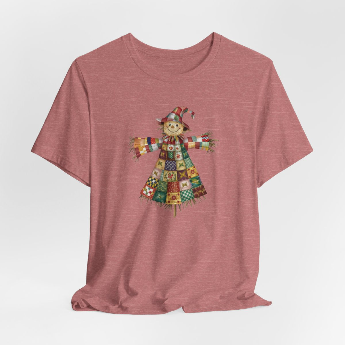 A Heather Mauve thanksgiving  quilting t-shirt featuring a smiling scarecrow made entirely of colorful quilt patterns. Wearing a patchwork hat and clothes with various quilt designs. Straw details on hands and neck. Cheerful autumn-themed illustration celebrating quilting craft.