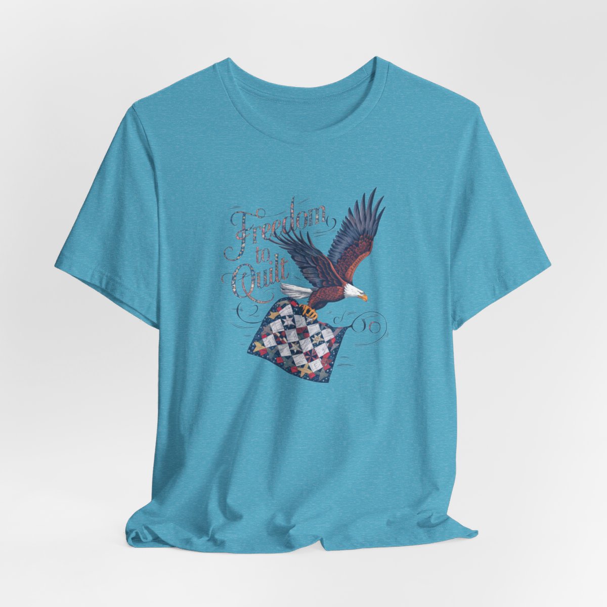 A Heather Aqua T-shirt with detailed illustration of a bald eagle in flight, carrying a patchwork quilt. Text reads "Freedom to Quilt" in an ornate, patriotic font.