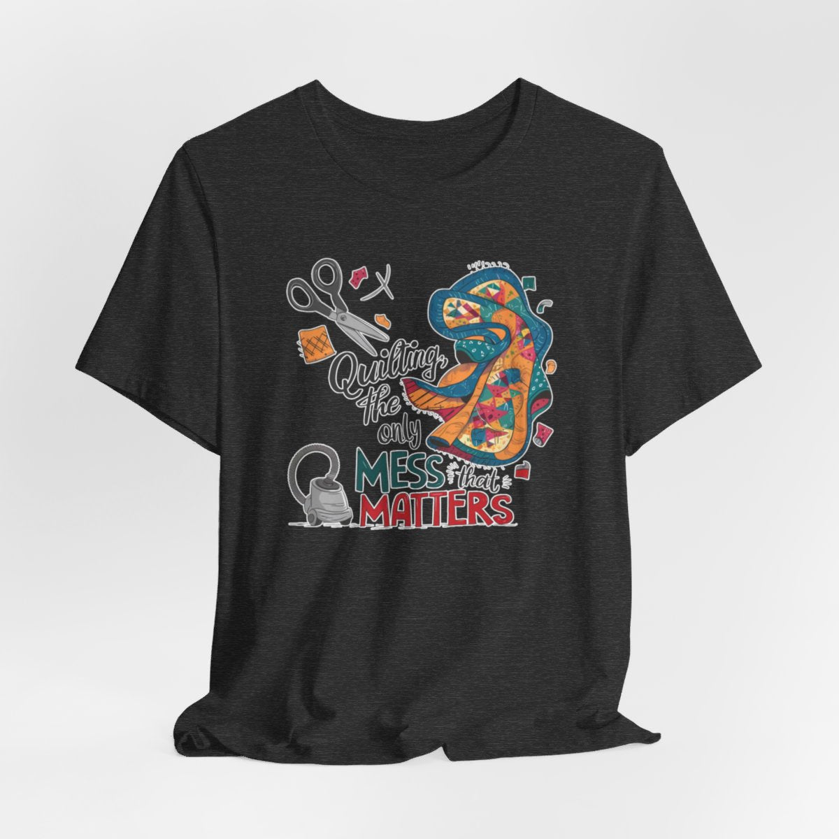 A Dark Grey Heather fun T-Shirt with the phrase Quilting, the Only Mess that Matters and illustrations of fabric scraps, scissors, and a vacuum cleaner. Colorful quilt patterns swirl around the text, creating a playful and relatable design for quilting enthusiasts.