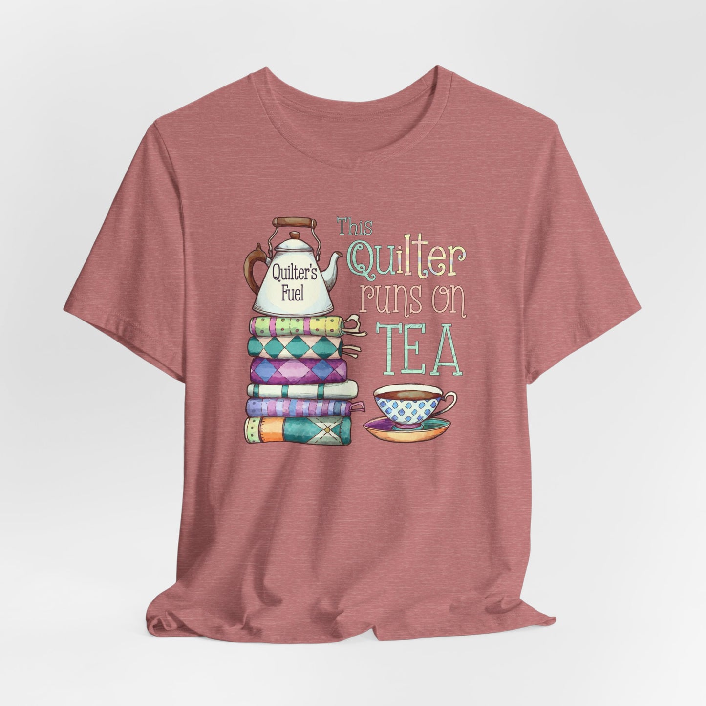 A Heather Mauve t-shirt featuring a whimsical design of a stack of patchwork quilts with a teapot labeled 'Quilter's Fuel' and the phrase 'This Quilter Runs on Tea,' ideal for tea-loving quilters.