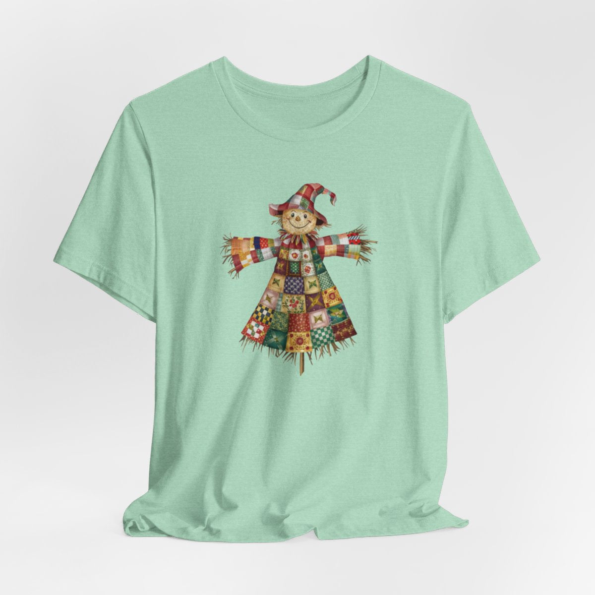 A Heather Mint thanksgiving  quilting t-shirt featuring a smiling scarecrow made entirely of colorful quilt patterns. Wearing a patchwork hat and clothes with various quilt designs. Straw details on hands and neck. Cheerful autumn-themed illustration celebrating quilting craft.