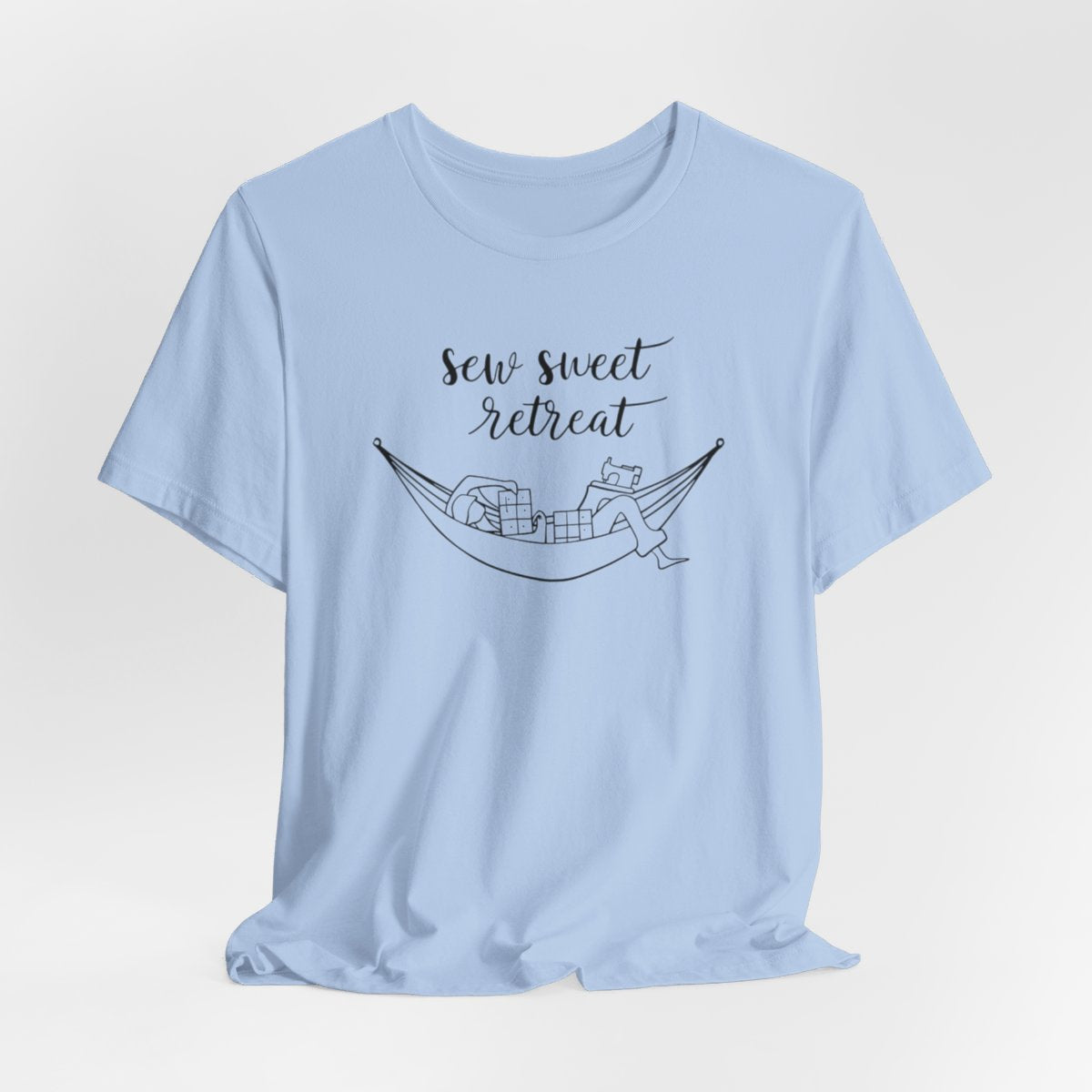 A Baby Blue T-Shirt with a black and white illustration of a hammock filled with quilting supplies like fabric squares and a sewing machine. Text above reads "Sew Sweet Retreat" in a flowing script font.